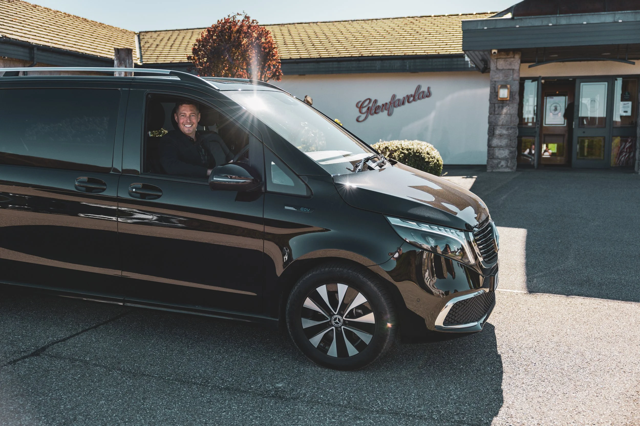 A Chauffeur smiling inside a black Mercedes-Benz electric vehicle parked outside a building with a sign that says 'Glenfarclas'.