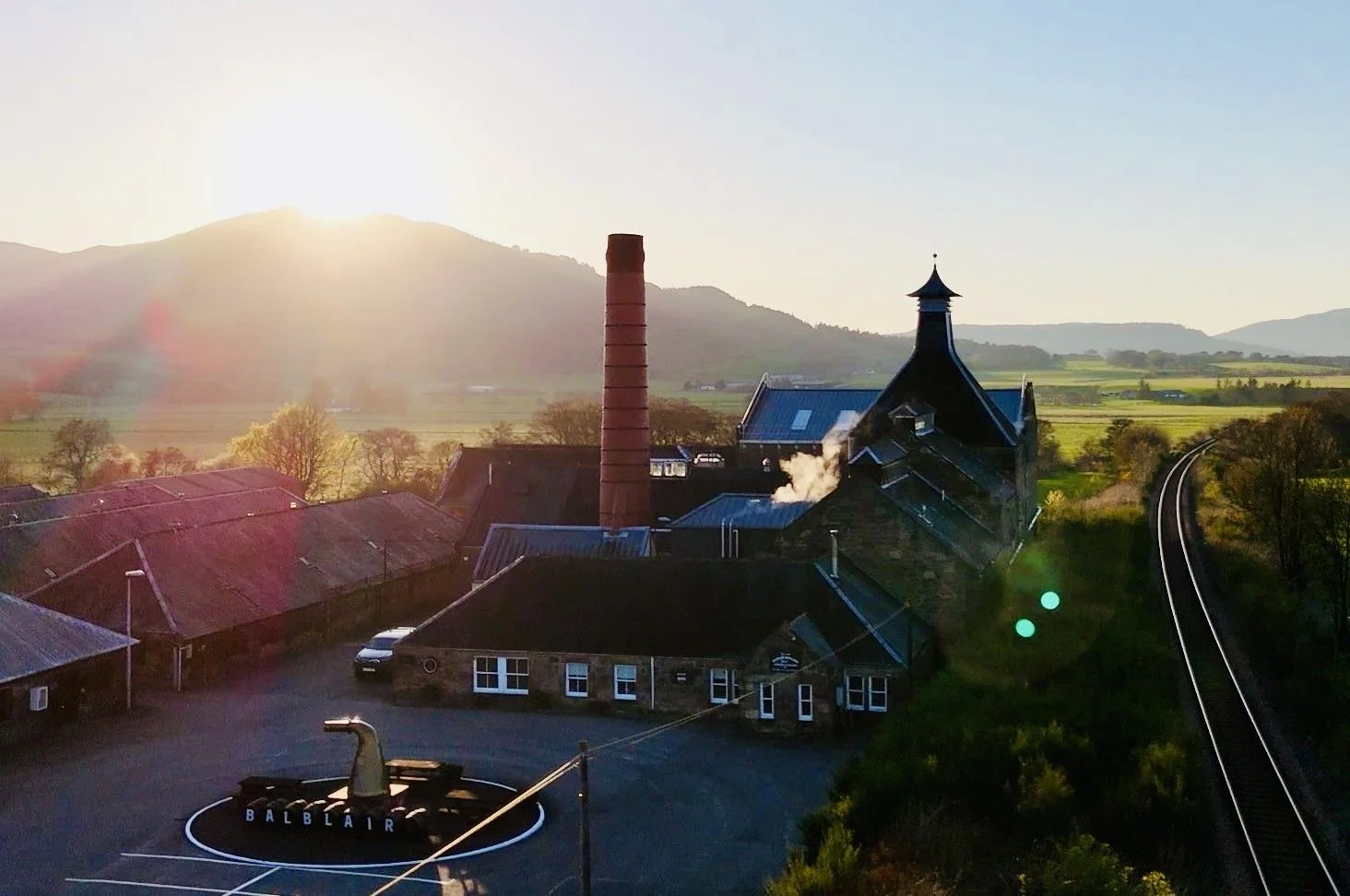 A rural landscape at sunset with a Distillery, a tall chimney, and rolling hills in the background. The Distillery has a blue roof and an ornate roof. There is a roundabout with a duck sculpture and the word "BALBLAIR" in front of it.