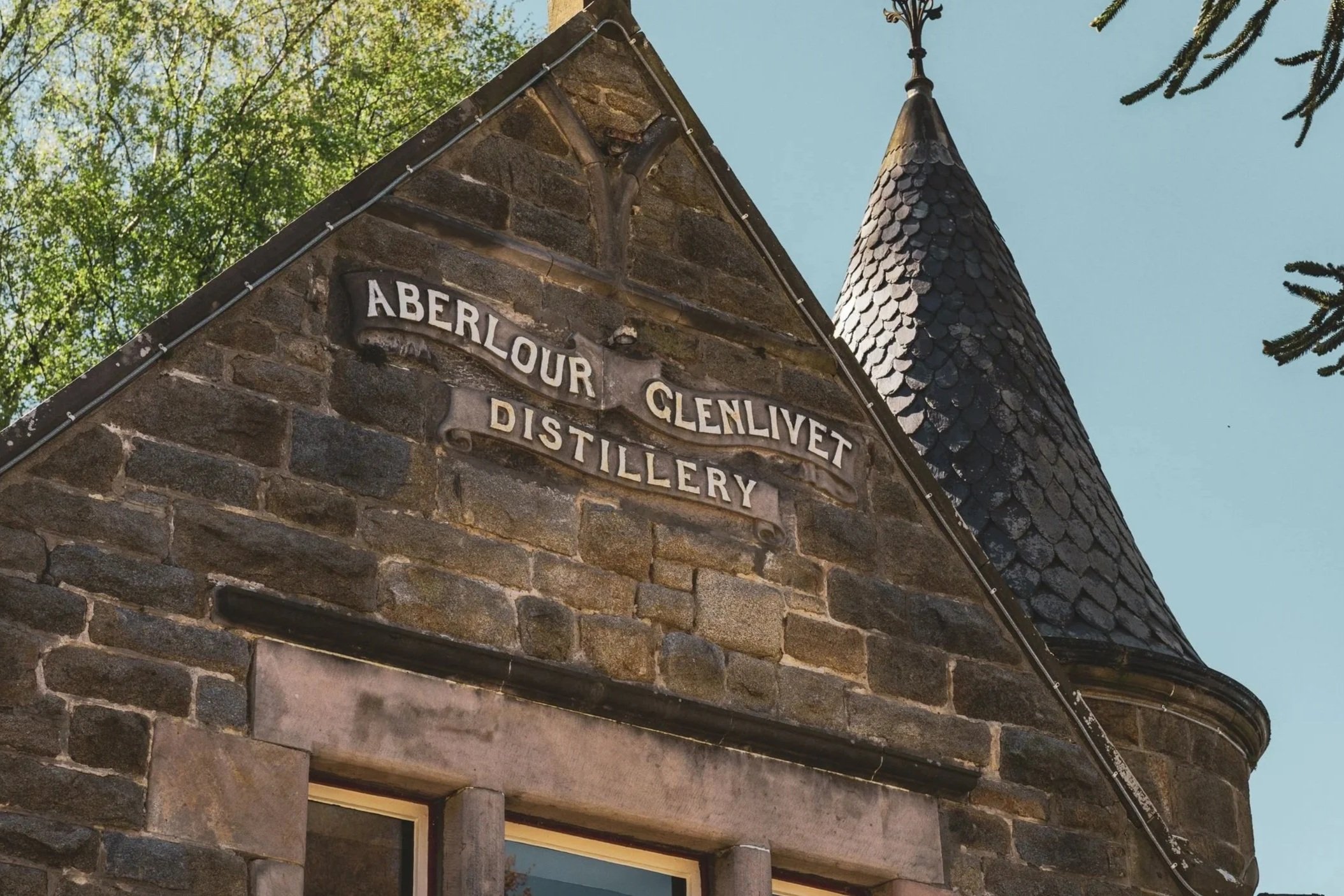 Close-up of a stone building with a sign that reads 'Aberlour Glenlivet Distillery' and a steep, shingled roof with a decorative spire, surrounded by green trees and a clear blue sky.