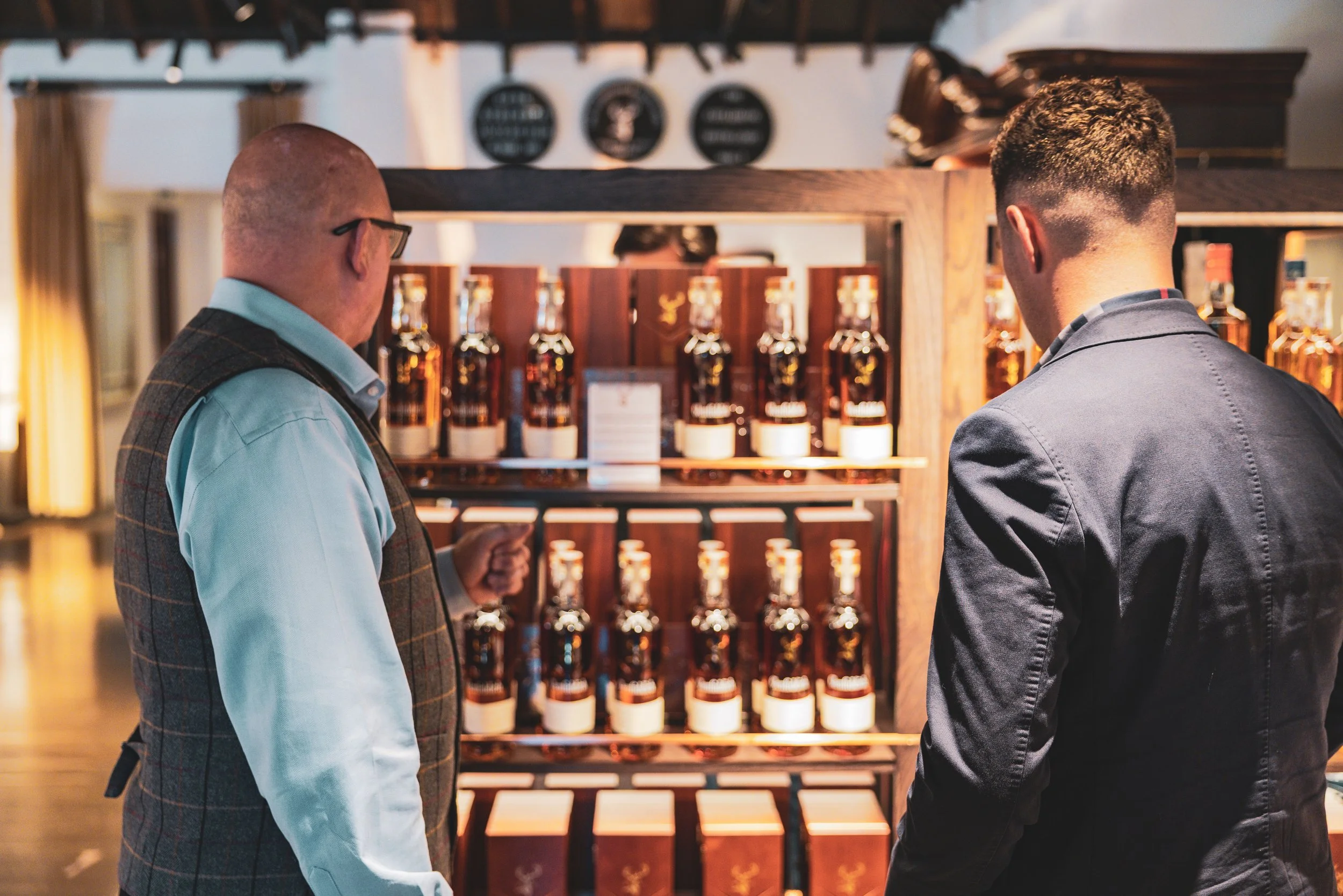 Two men are standing in front of a shelf of whisky bottles, facing each other in a dimly lit room.