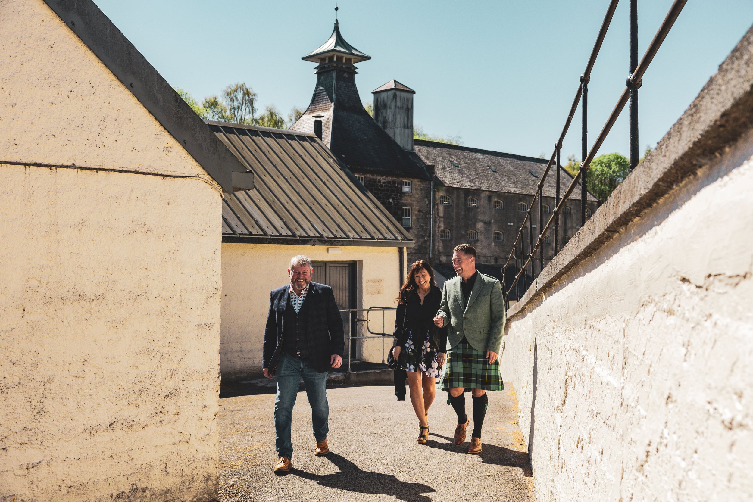 Three people, two men and one woman, walking outdoors between stone walls with a historic building and blue sky in the background. The man on the right is wearing traditional Scottish attire including a kilt and jacket.