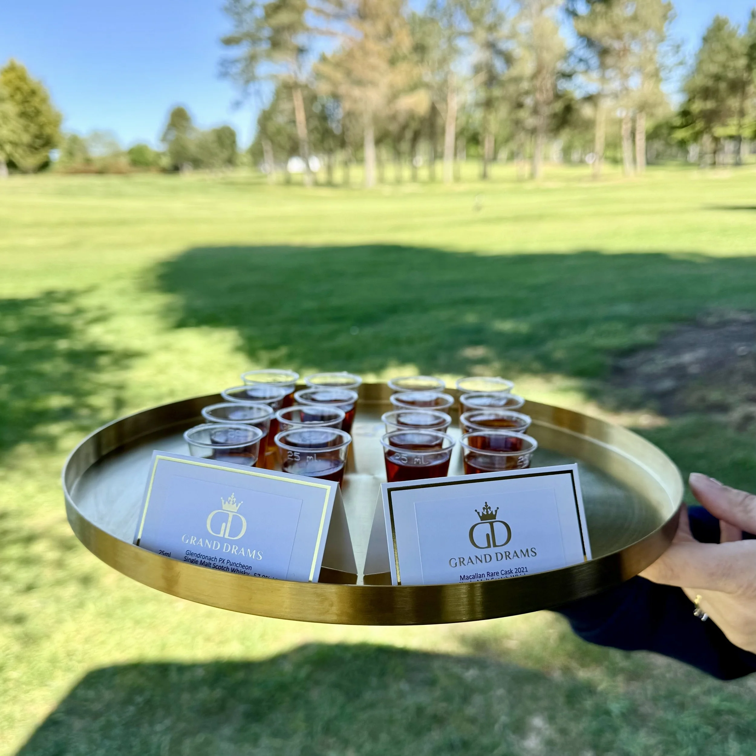 A person holding a gold tray with small glasses of whisky and labeled cards on a grassy outdoor area.