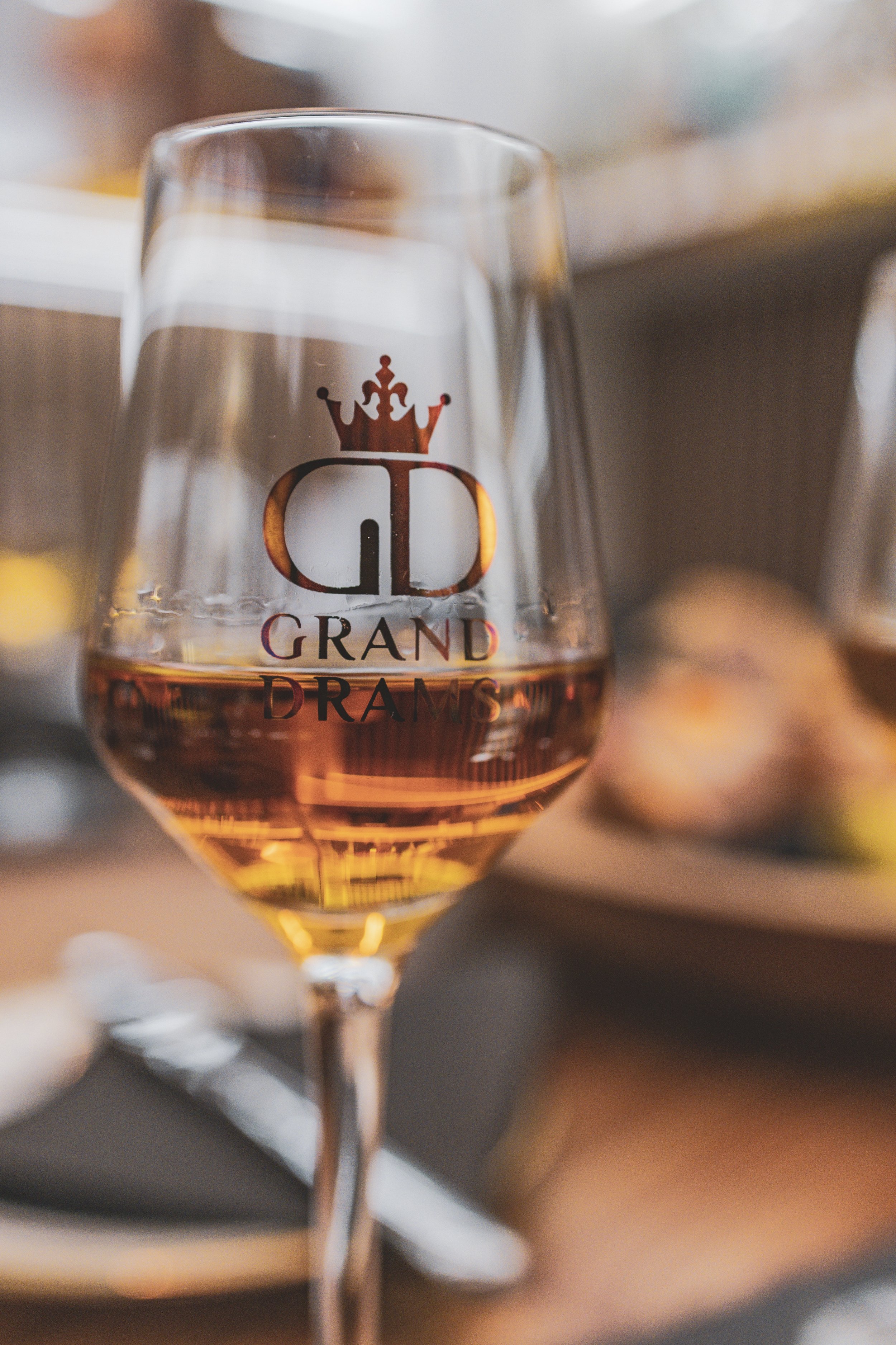 A wine glass with a logo featuring a crown and the text "Grand Dams" filled with rosé wine, placed on a table with utensils and a blurred plate of food in the background.
