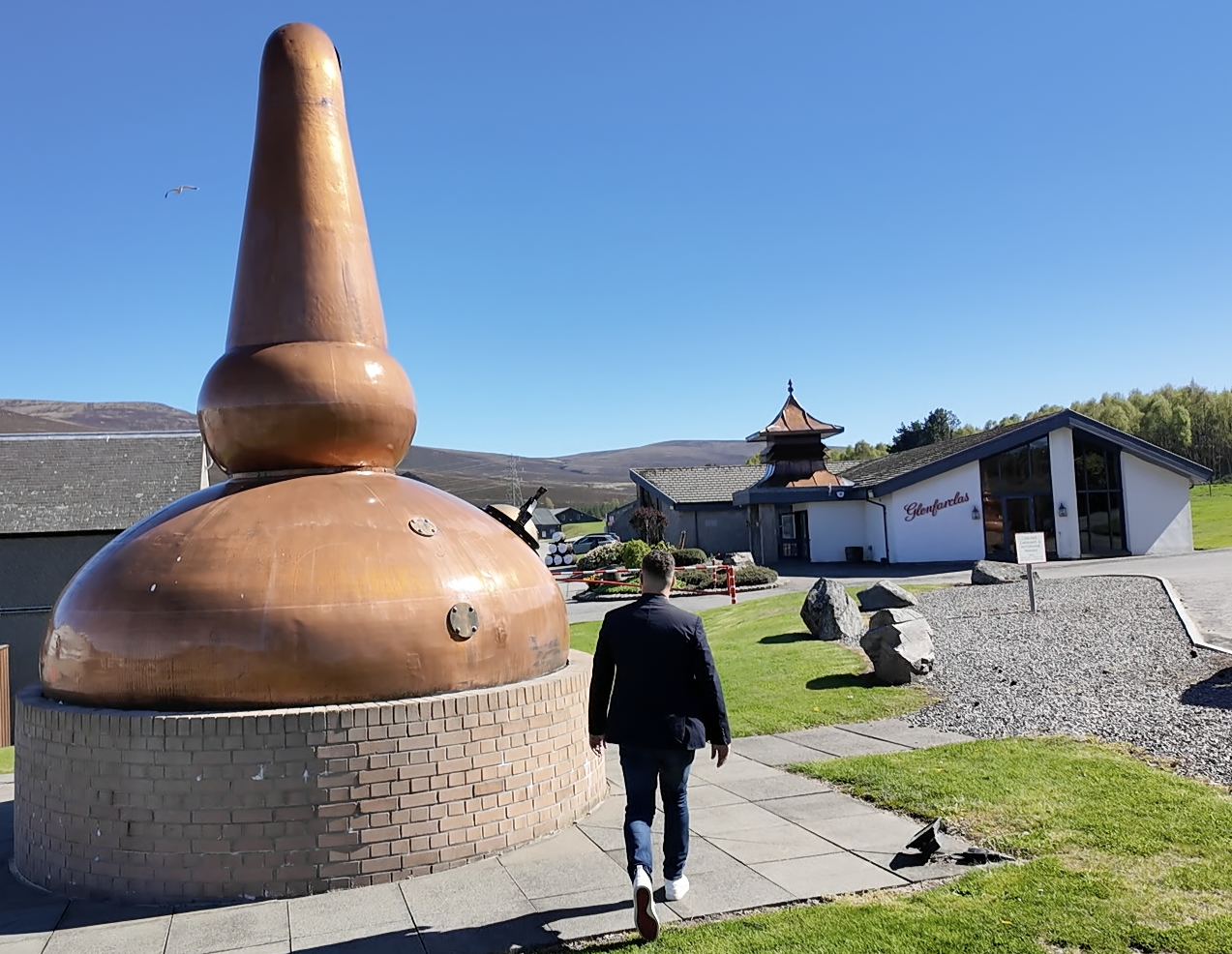 A man walking past a large copper still resembling a traditional Scottish bagpipe outside a modern distillery building with a sign that reads 'Glenfarclas' under a clear blue sky.