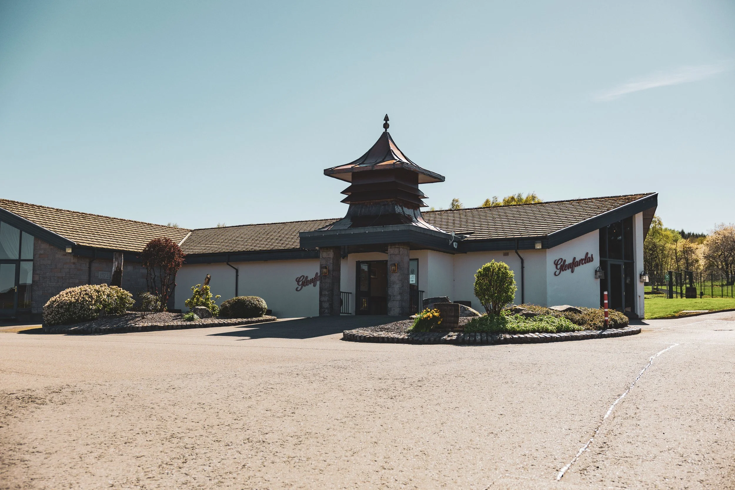 A building with a traditional Asian-style pagoda roof and modern architecture, surrounded by landscaped plants, with a parking lot in front and a clear blue sky above.