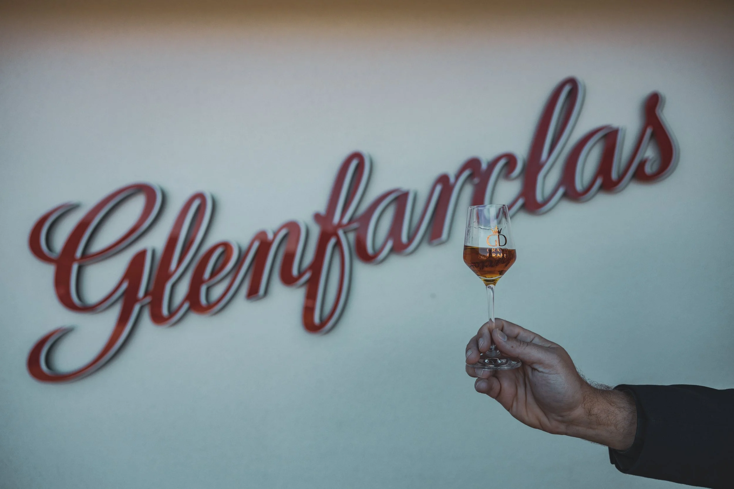 A person holding a wine glass filled with a pinkish beverage in front of a wall with a large red cursive sign that says "Celebramos."