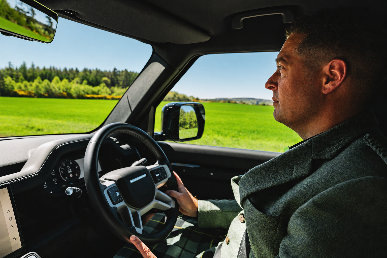 A man in a kilt driving a vehicle through a rural landscape with green fields and trees under a clear blue sky.