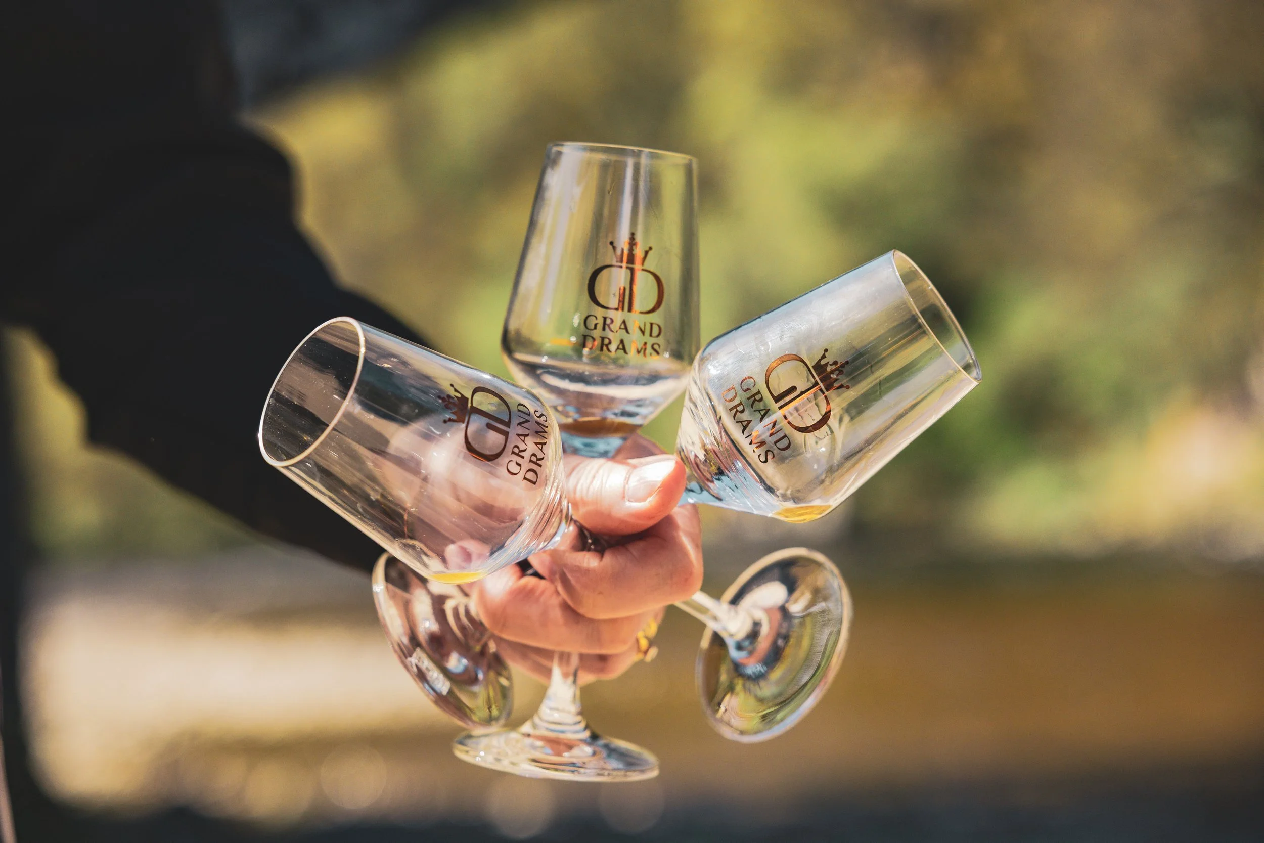 Multiple empty wine glasses with the logo 'Grand Drams' held by a person outdoors, with a blurred background of trees and sunlight.