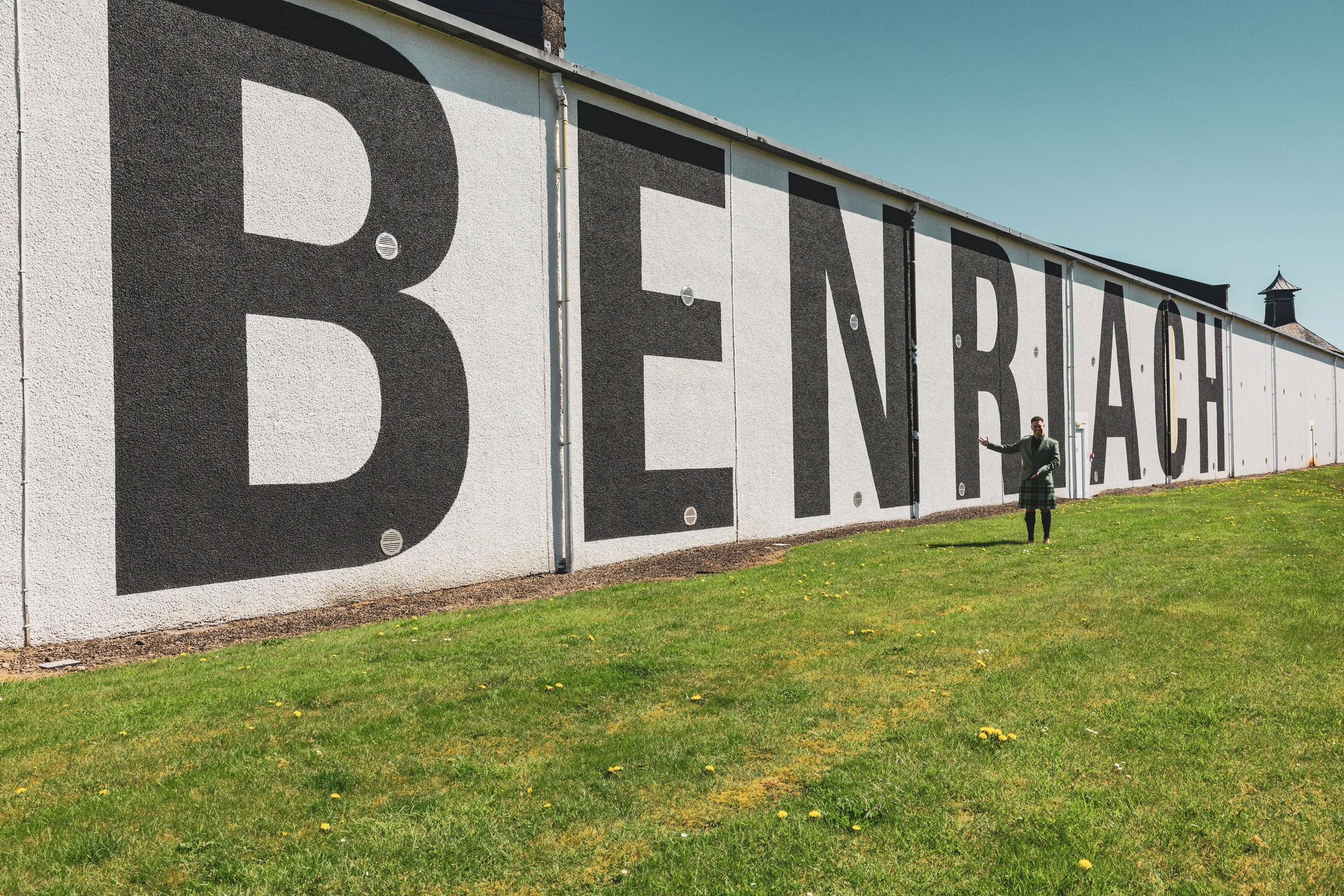 A woman standing in front of a wall with large black text that reads 'BEREK'. She is holding her hand out towards the wall. The wall is part of a building with a small structure or tower visible in the background. The grass in the foreground is green
