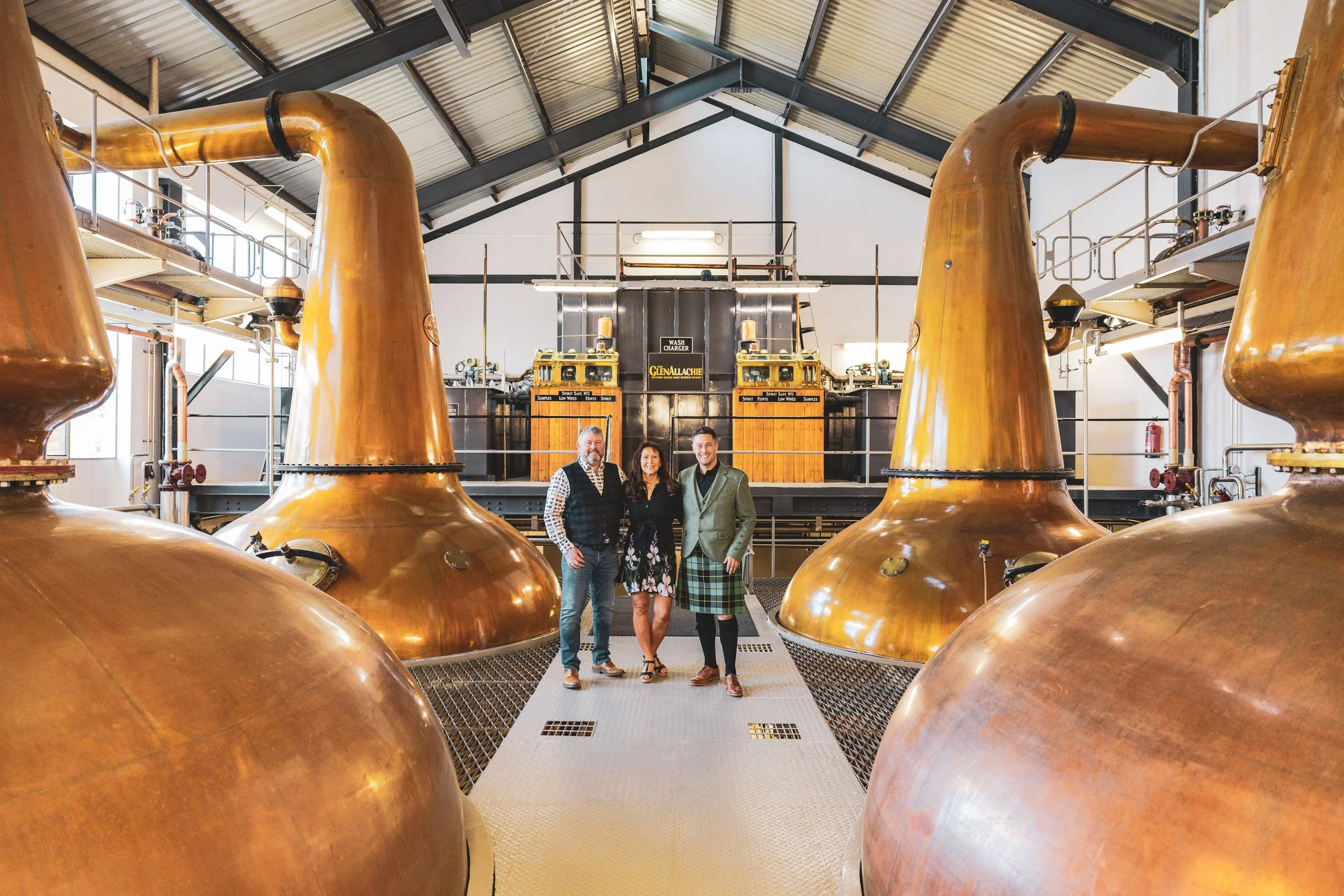 Three people standing inside a whiskey distillery with large copper stills around them. The stills are shiny and copper-colored, and the building has a high ceiling with metal roofing. The group is smiling and dressed in business casual attire.