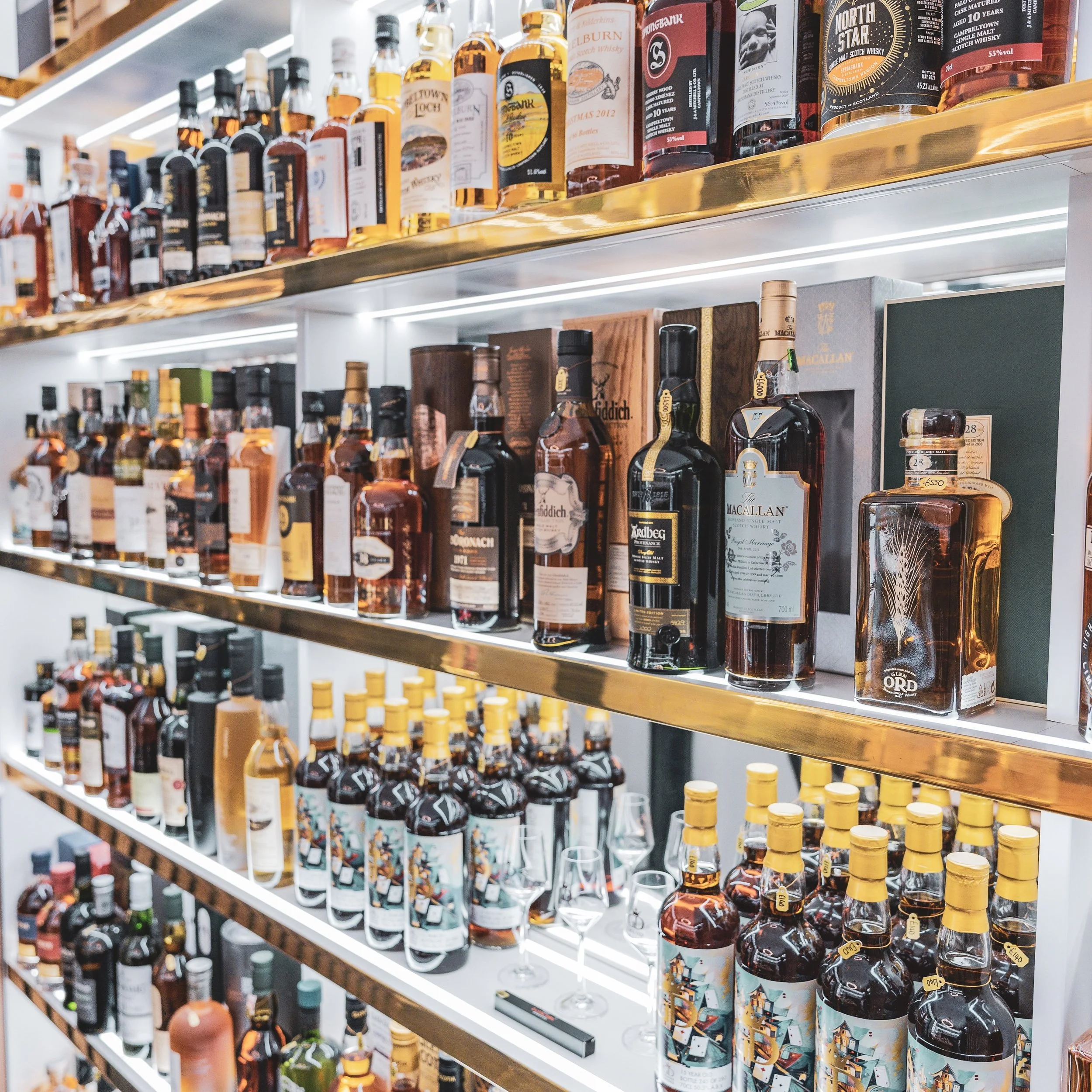 A store shelf stocked with various bottles of whiskey and spirits, arranged on illuminated glass shelves with gold-colored edges.