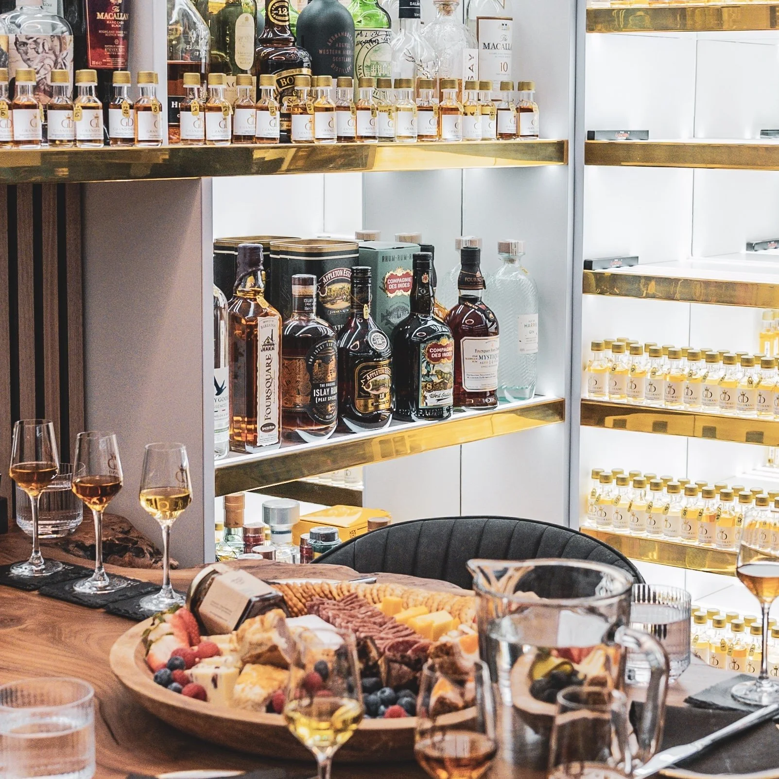 A bar or dining area with a wooden table set with glasses of white wine, a cheese and charcuterie platter with berries, and beverages on shelves of liquor bottles, including whiskey and other spirits.