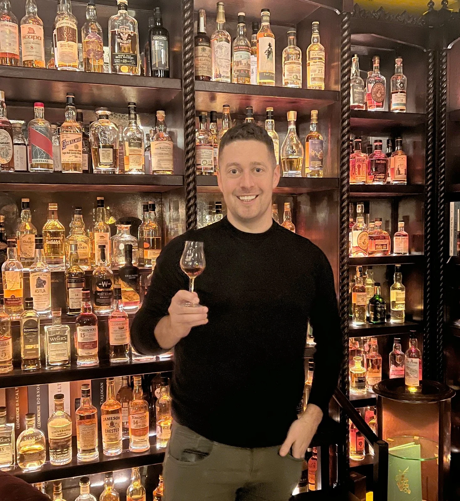 A man smiling and holding a glass of whiskey in front of a well-stocked bar with multiple bottles of liquor