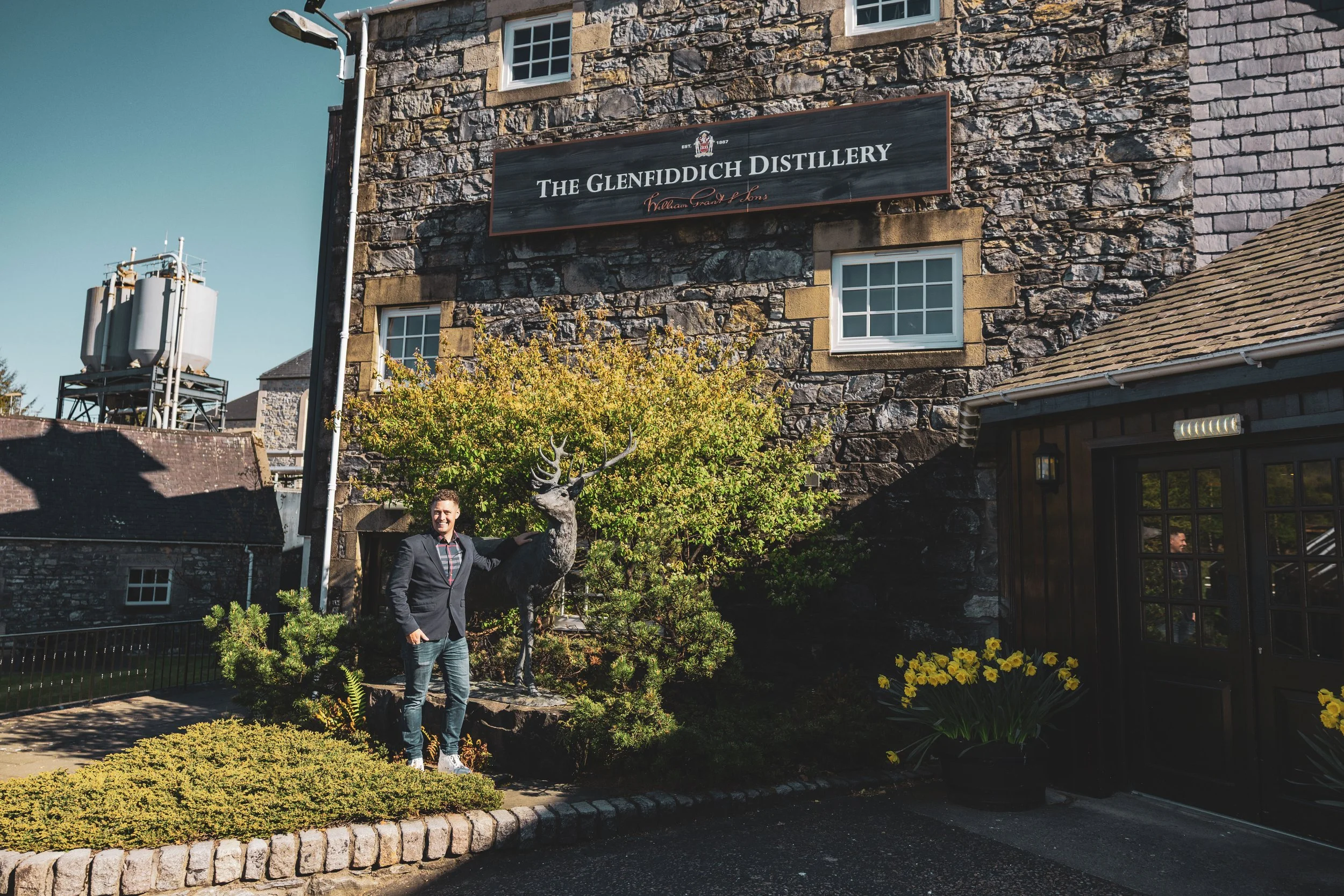 A man in a suit standing outdoors next to a deer statue in front of a building with a sign that reads "The Glenfiddich Distillery." There are plants and flowers in the garden area, and the day appears sunny.