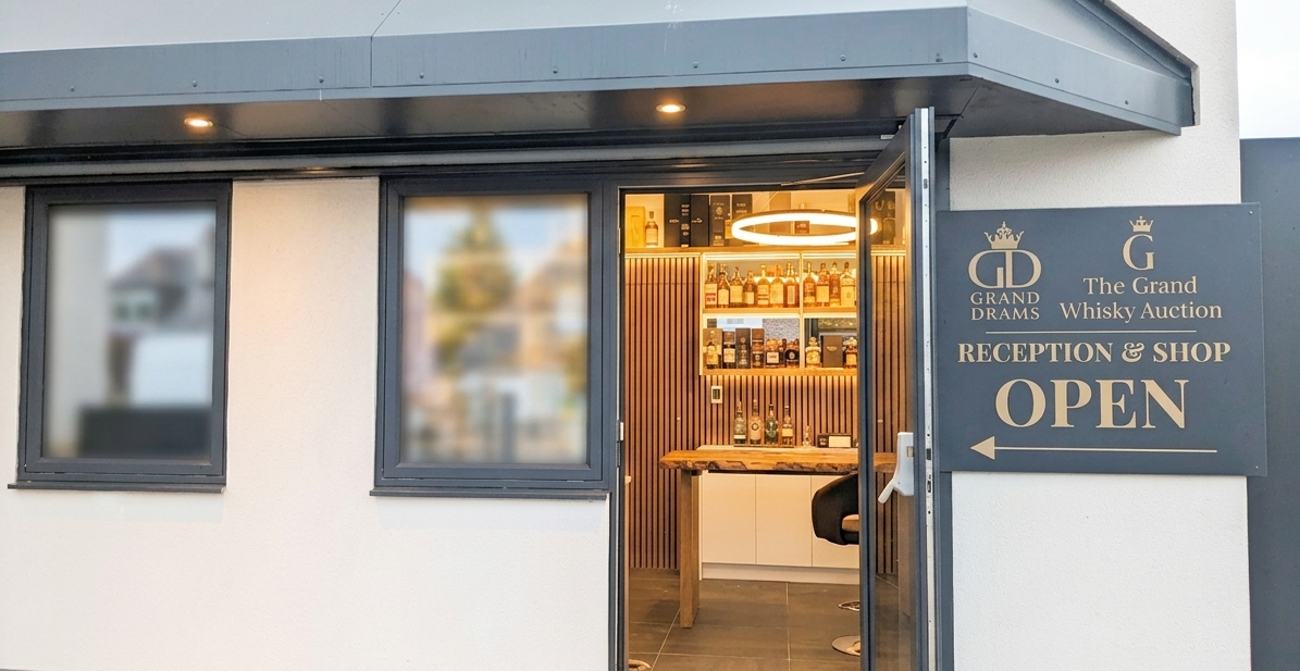 The entrance of a shop with a sign that reads 'Grand Drams, The Grand Whisky Auction, Reception & Shop, Open'. The shop has a door open showing a bar inside with bottles of whisky on shelves.