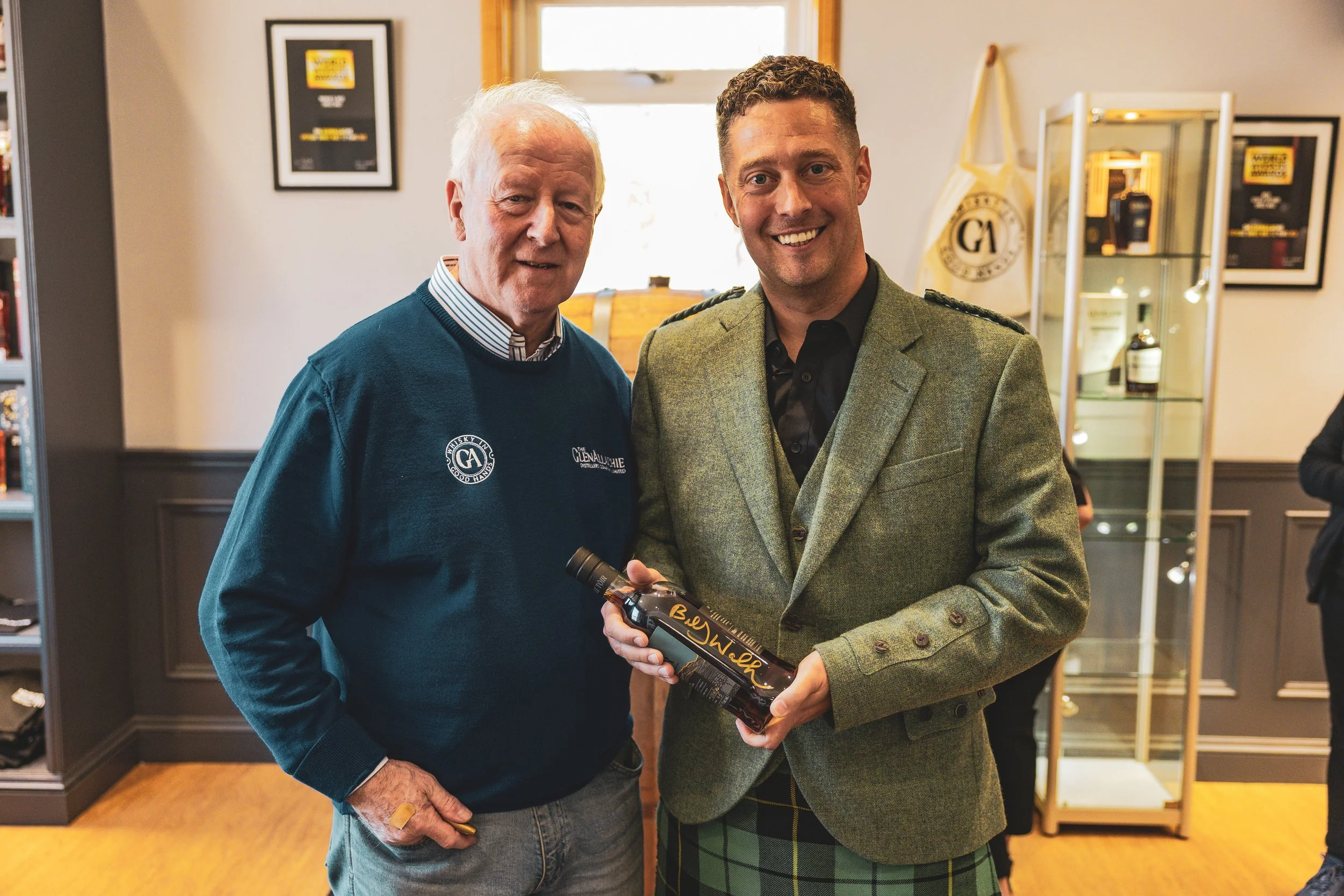 Two men smiling, standing indoors near a display case with bottles and framed pictures on the wall. The older man on the left is wearing a navy blue sweater with logos, and the younger man on the right is wearing a green plaid suit and holding a bott