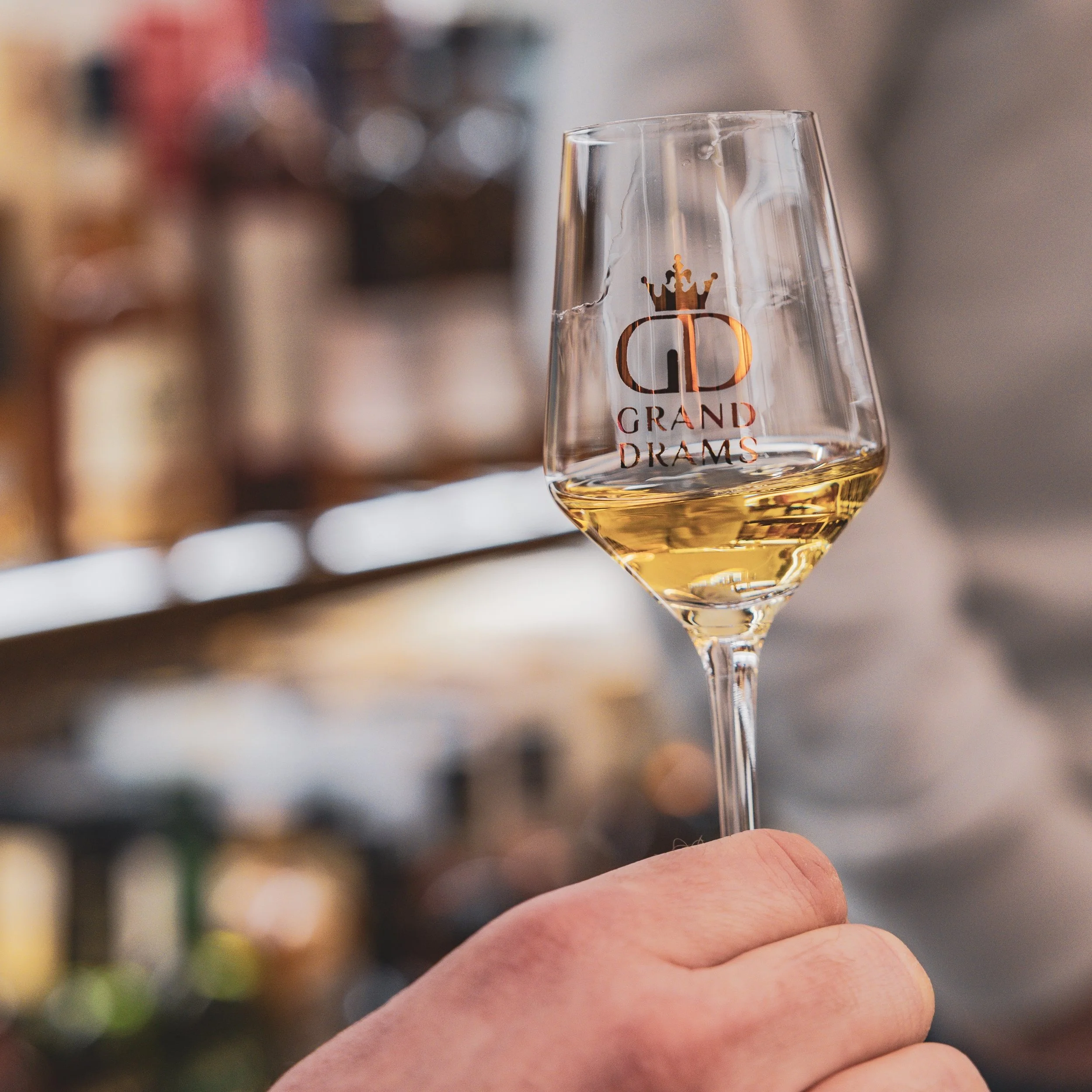 A hand holding a glass of white wine with a logo and text 'Grand Drams' on the glass, blurred shelves in the background.