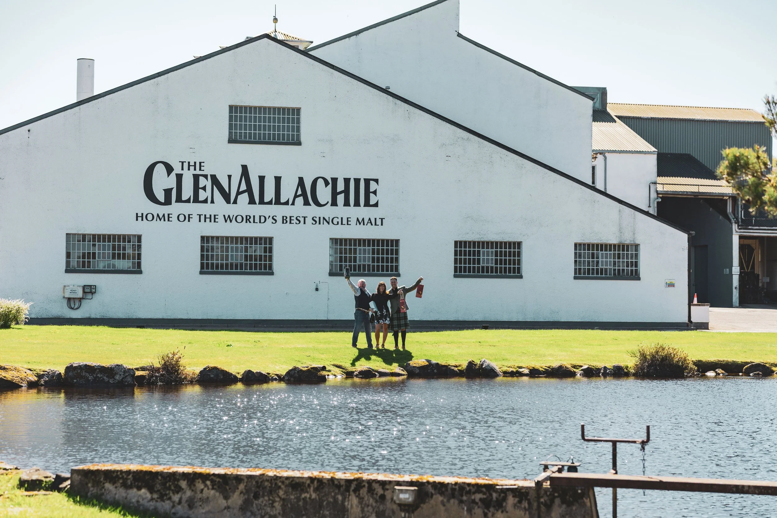 A white building with black lettering that reads "The Glentallachie, Home of the World's Best Single Malt". There are three people standing on the grass in front of the building, posing for a photo. A body of water is in the foreground.
