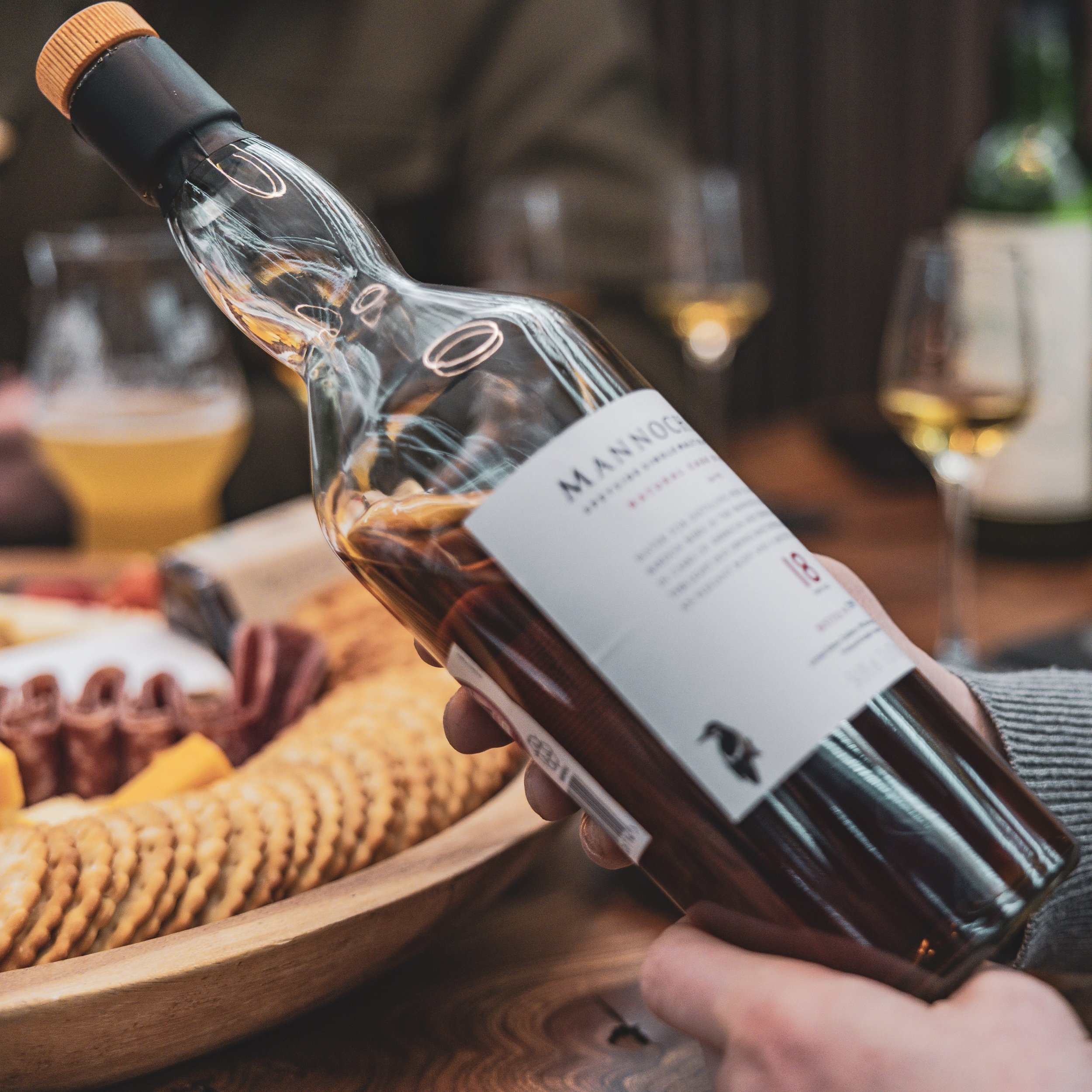 Hand holding a bottle of whisky pouring into a glass at a table with a charcuterie board in the background.