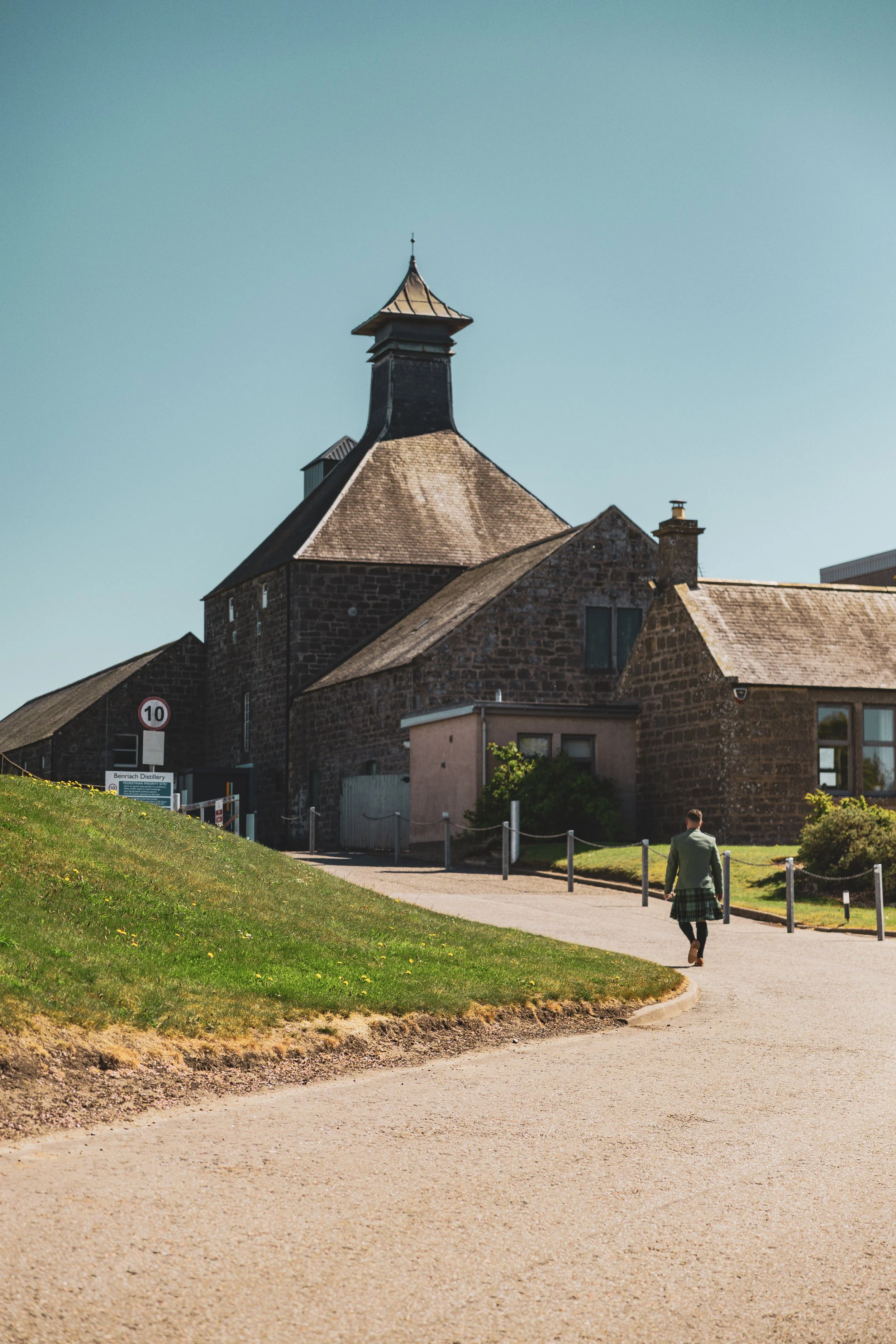 A man dressed in a traditional Scottish kilt walking on a paved pathway near buildings made of dark stone, with a grassy area on the left and a clear blue sky overhead.