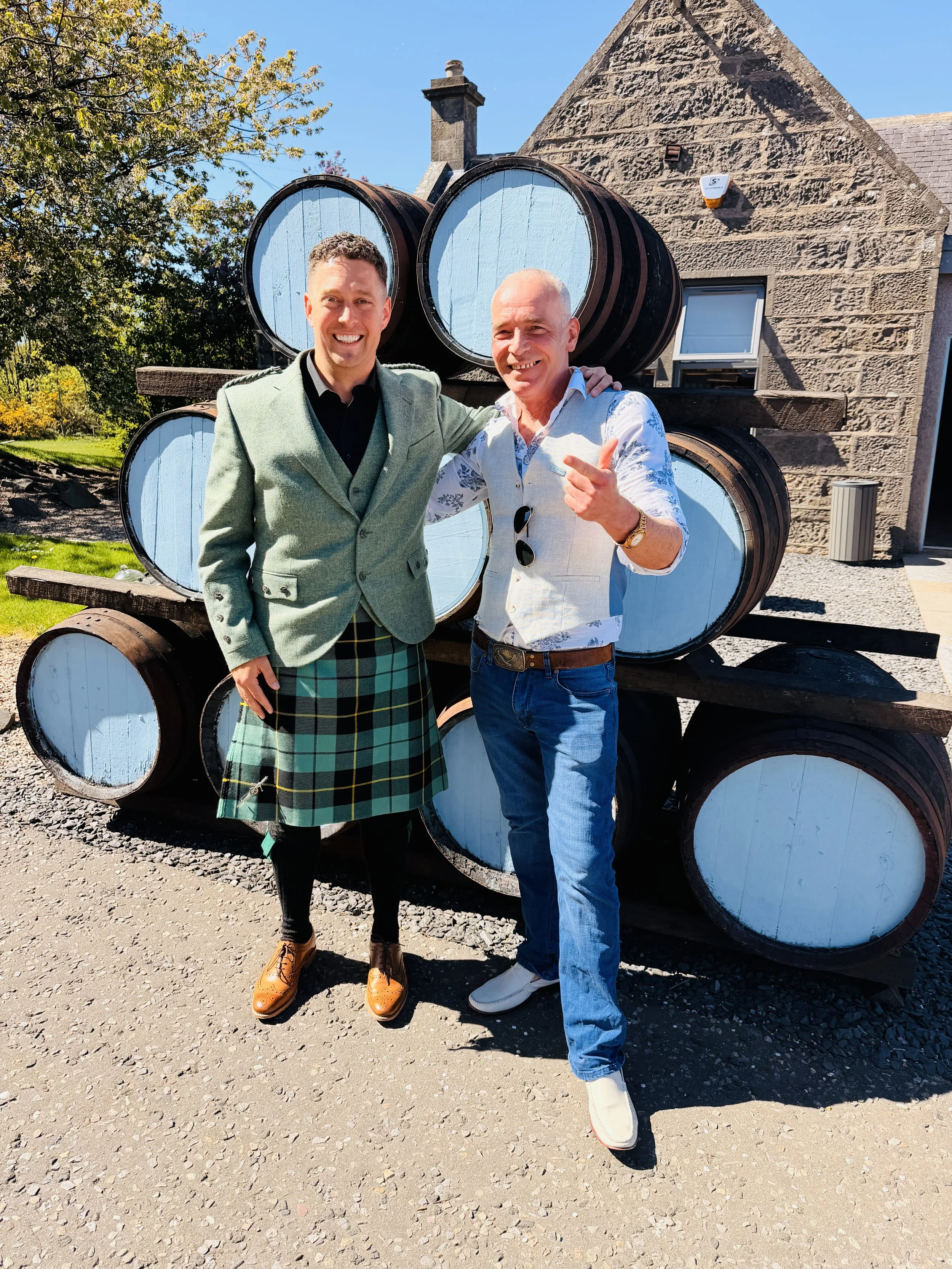 Two men standing outdoors in front of stacked barrels, smiling and posing for the photo. One man is wearing a green blazer and tartan kilt, the other is dressed in a white shirt with blue jeans, pointing at the camera. The background features a stone building on a sunny day.
