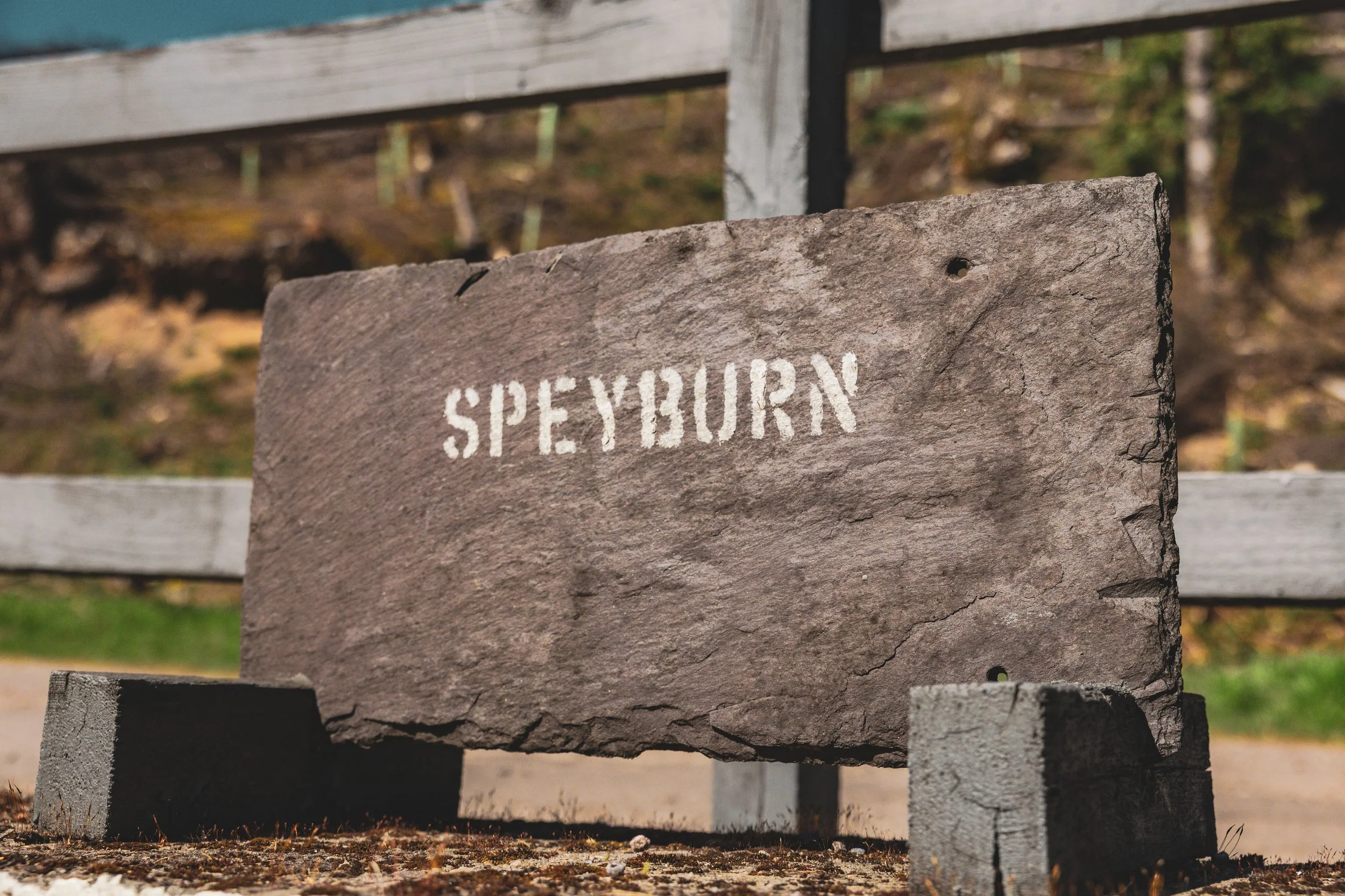 A weathered stone sign with white stenciled text reading 'SPEYSBUR' and part of the connected word missing, mounted on a wooden post outdoors, with blurred trees and foliage in the background.