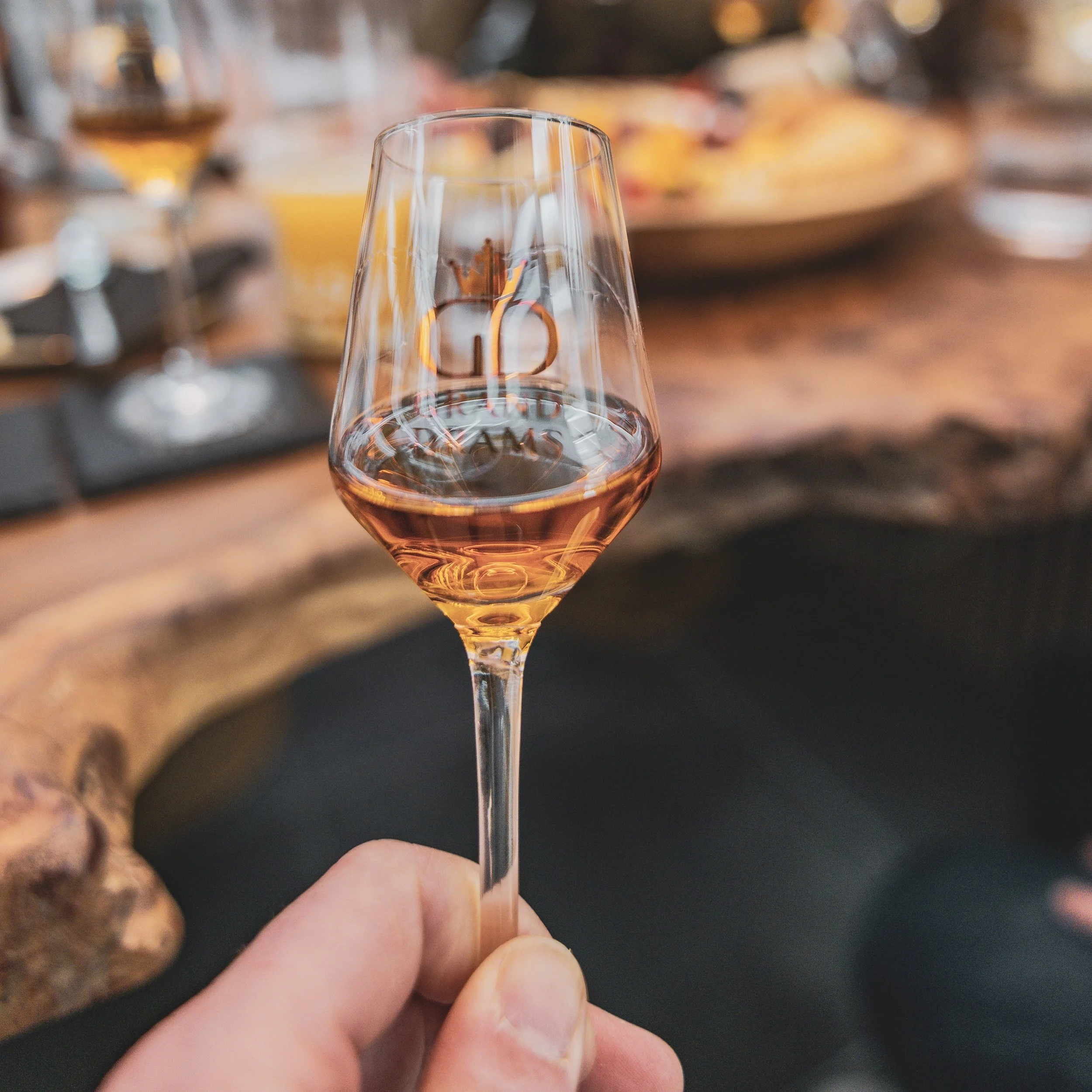 Close-up of a Grand Drams whisky glass held between fingers, with the letter 'GD' embossed on it, against a blurred background of food and drink on a table.