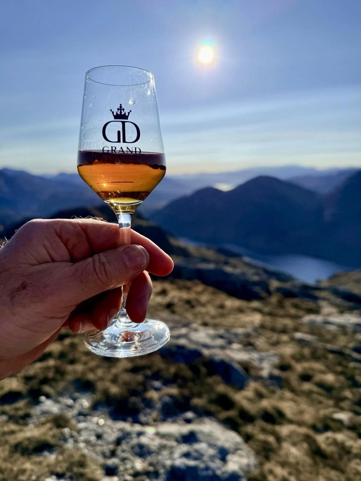 A hand holding a whisky glass with a logo that says 'Grand Drams' and a crown above a stylised 'GD' logo, against a Scottish mountain landscape under a bright sun in the sky.