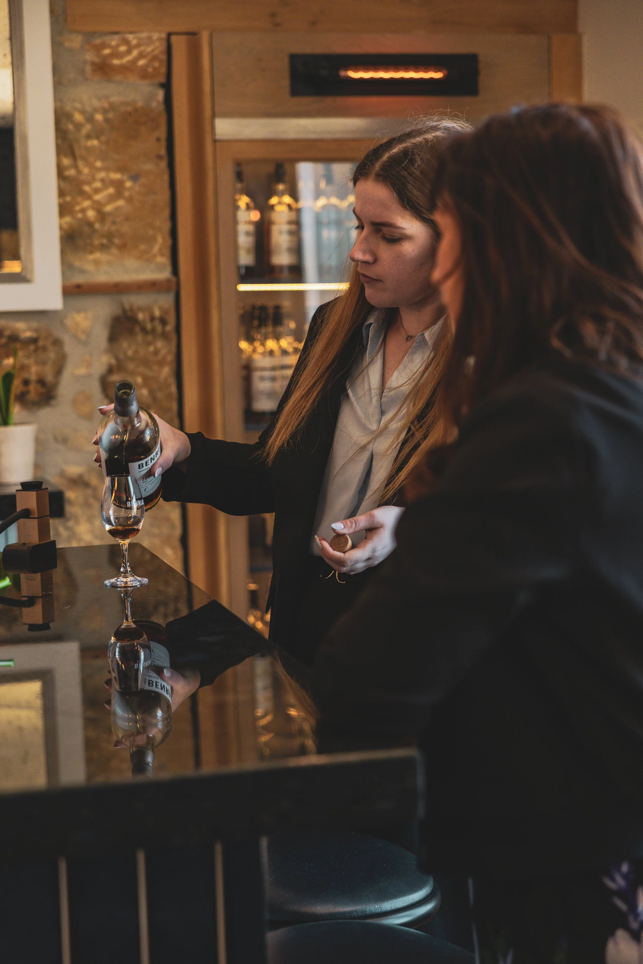 A woman with long brown hair pours a bottle of whisky into a Grand Drams glass at a bar. Another woman with dark hair is in the foreground, partially visible.
