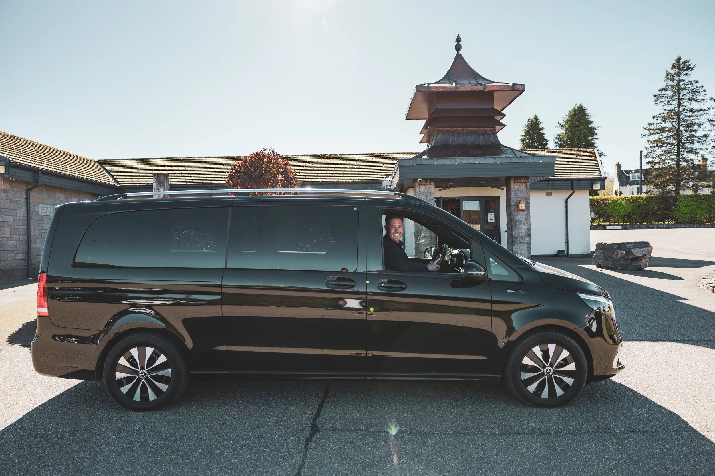 A man in a black shirt smiles inside a dark black van parked in front of a distillery entrance.