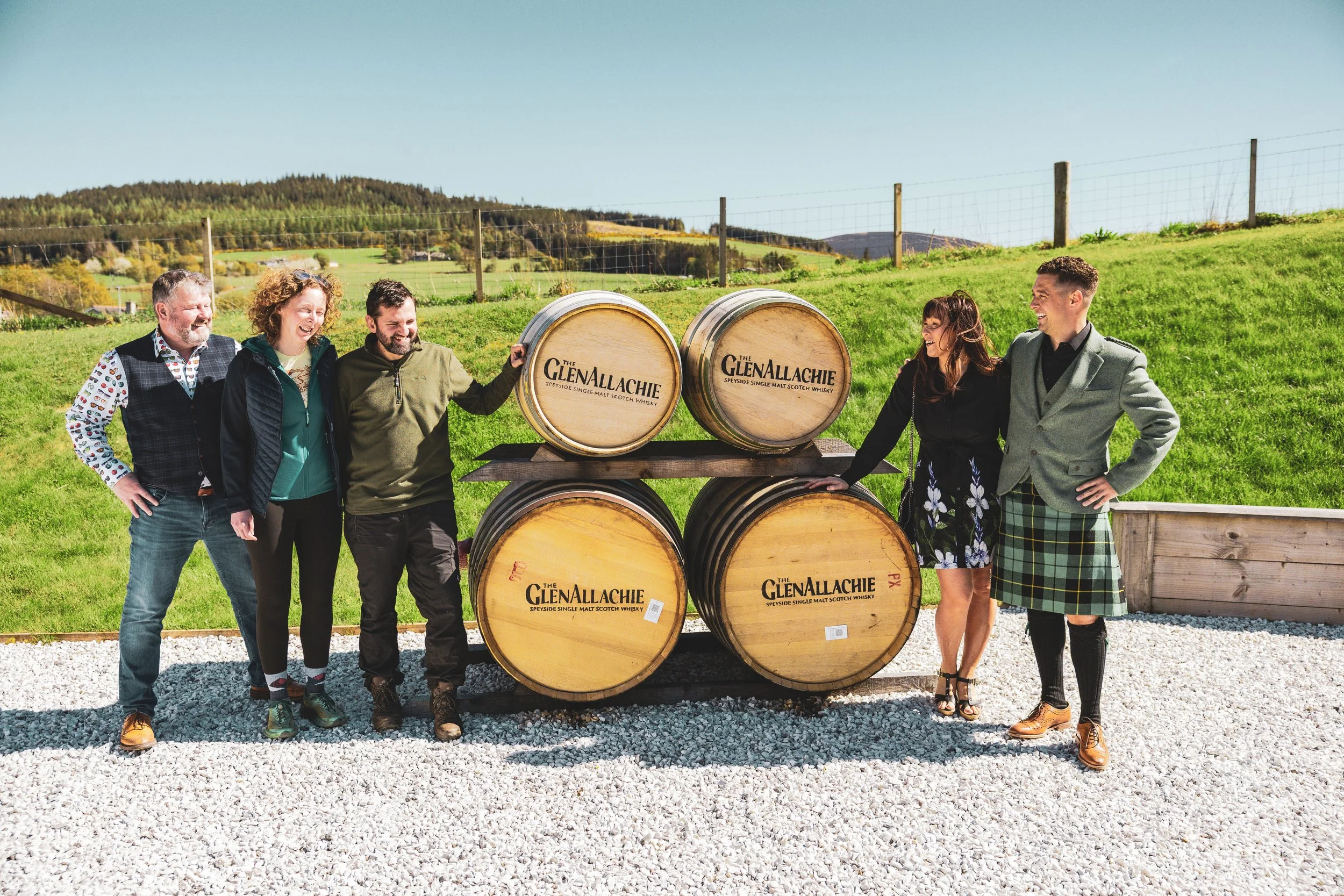 Group of six people standing outdoors with whiskey barrels labeled The GlenAllachie, smiling and laughing in a scenic rural setting with green fields and hills.