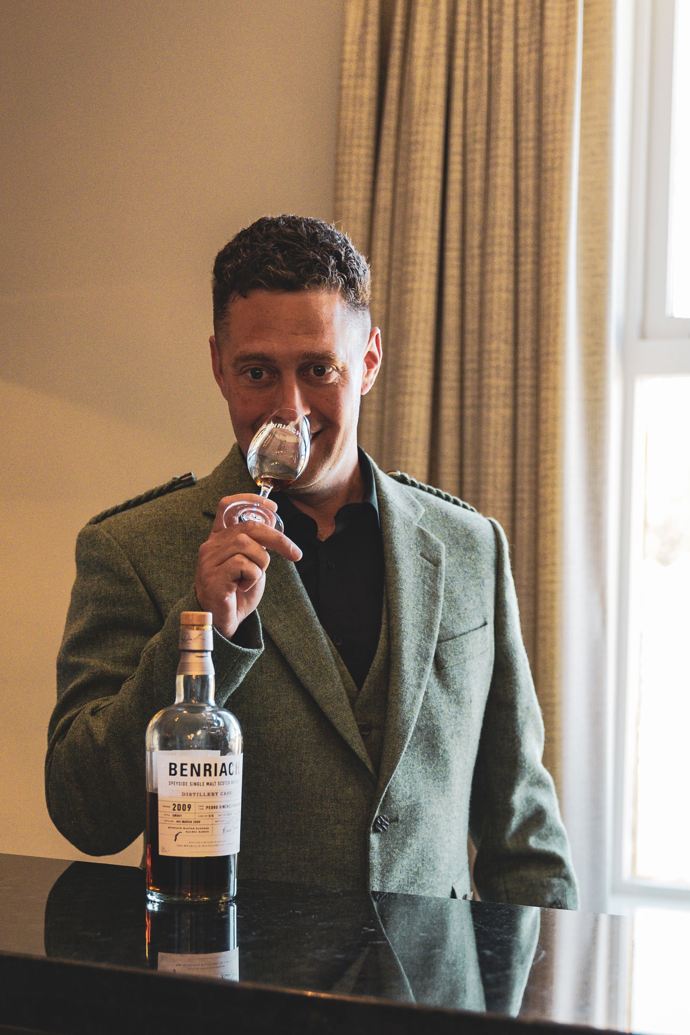 A man in a gray blazer drinking whiskey from a glass, with a bottle of Benriach whiskey on the counter in front of him, in a room with beige walls and a window with beige curtains.