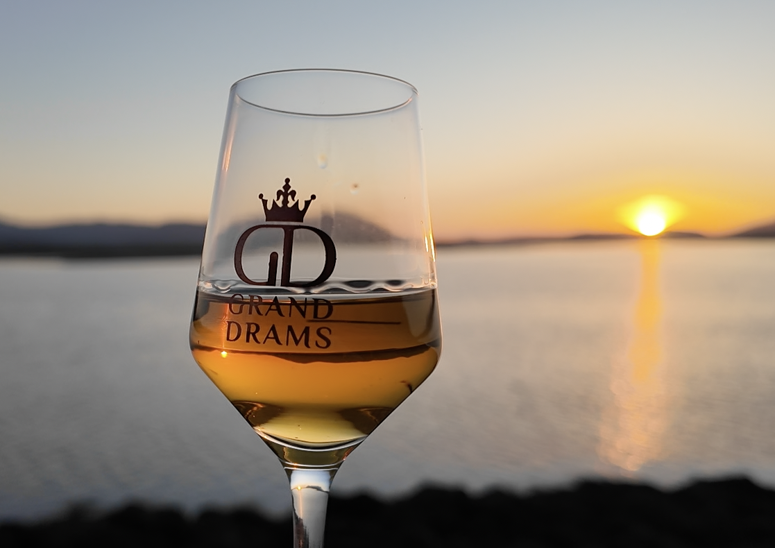 A glass of Scottish single malt whisky with the logo 'Grand Drams' and a crown, set against a sunset over a body of water.