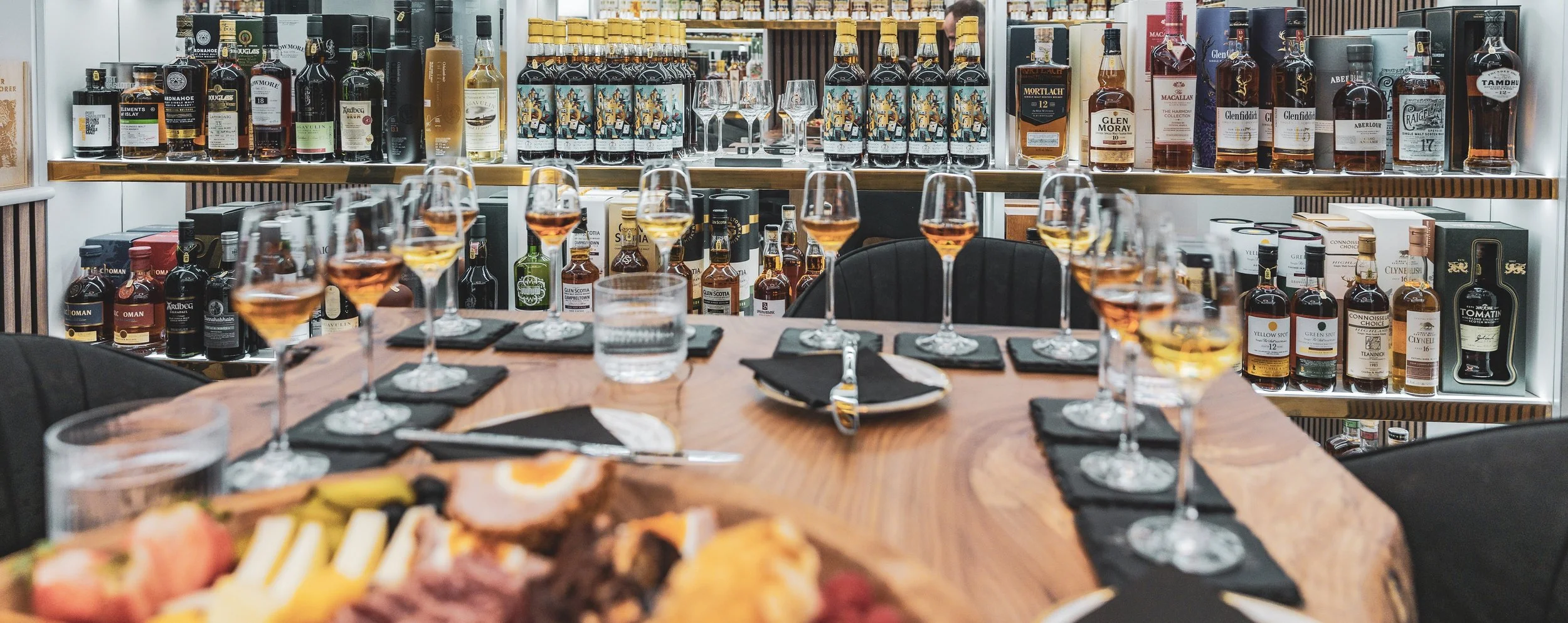 A table set for a whisky tasting event with glasses filled with whisky drams, and a charcuterie board in the foreground. In the background, shelves are stocked with various bottles of whiskey and other spirits.
