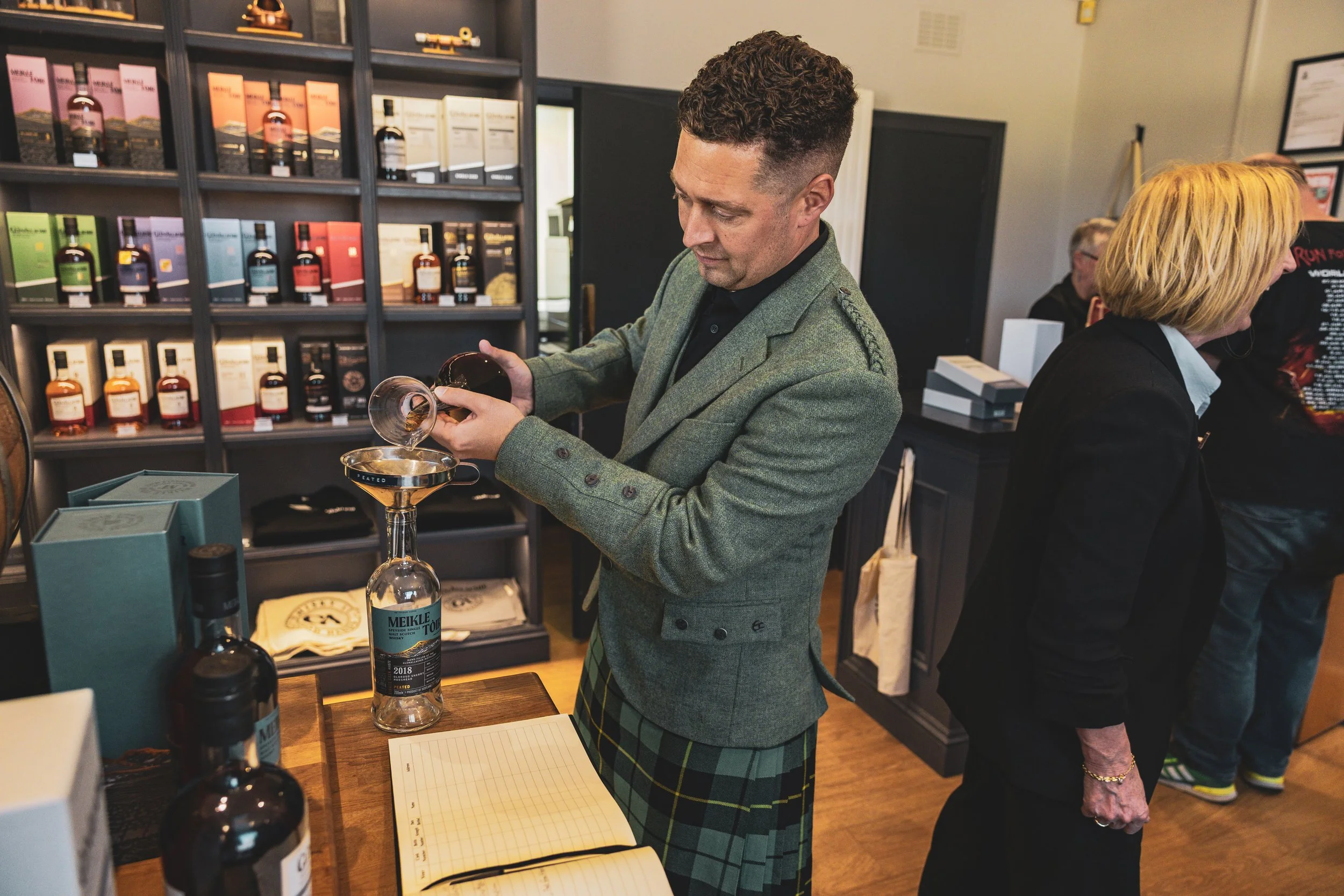 A man in a green blazer and plaid kilt is pouring whisky into a glass bottle at a tasting event, with shelves of bottles behind him and other people browsing in the background.