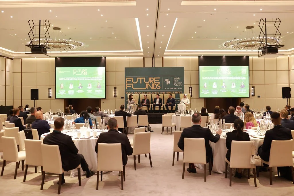 A conference event called 'Future Funds' held in a large, upscale ballroom with round tables, a panel of speakers on stage, and two large screens displaying the event title and details.