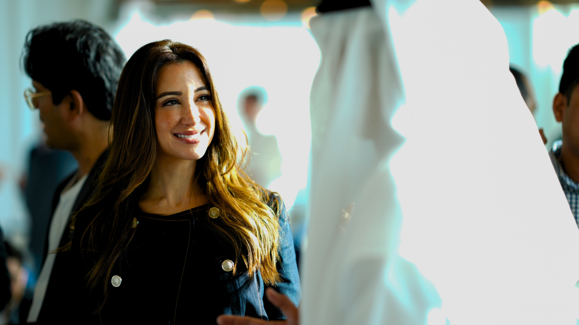 A woman with long brown hair, smiling, looking at a white dress on display in a busy clothing store or showroom with other people nearby.