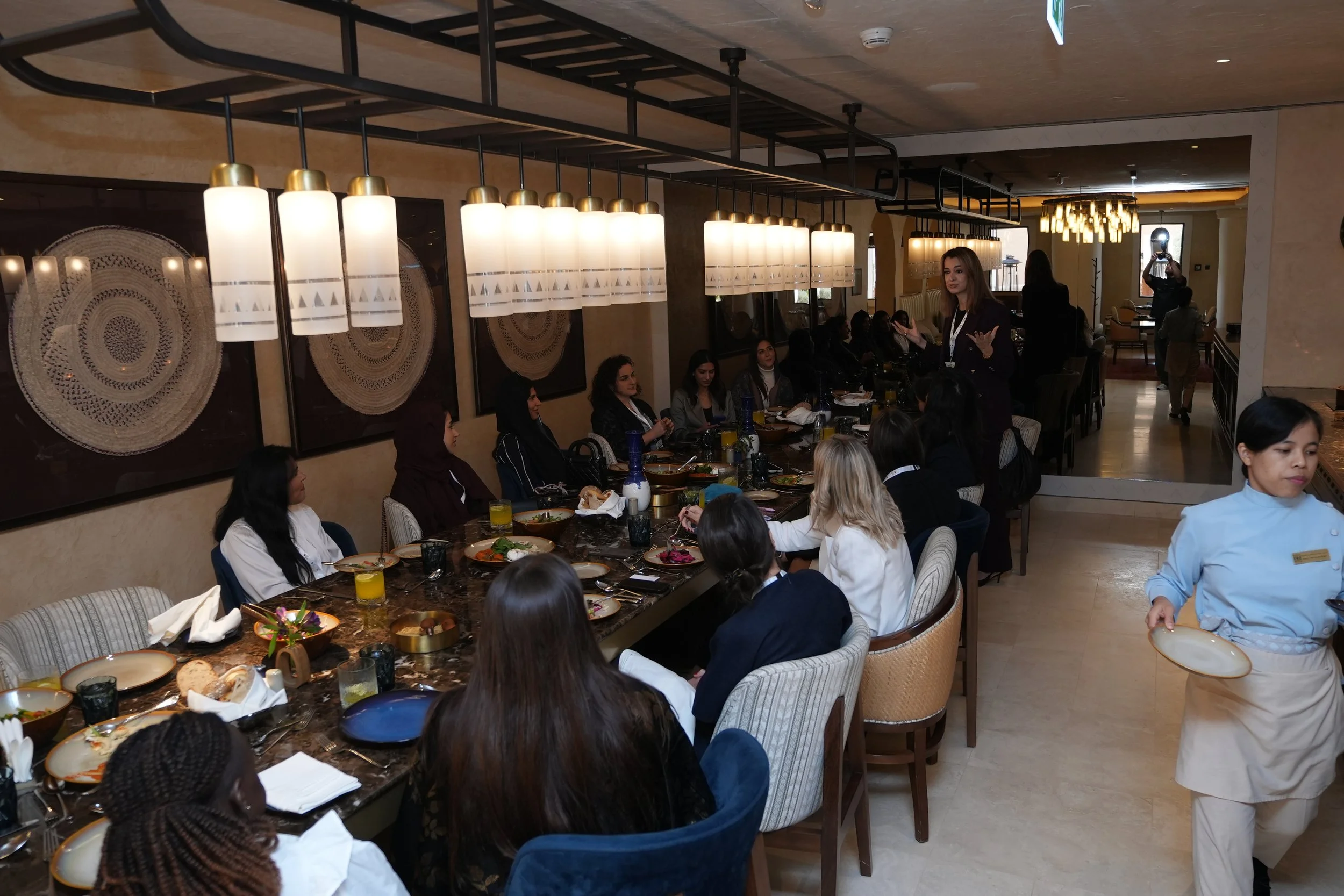 A diverse group of women sitting at a long dining table in a restaurant, with a woman standing and speaking to the group. The table is set with plates, dishes, drinks, and napkins. The restaurant has modern lighting fixtures and wall art.