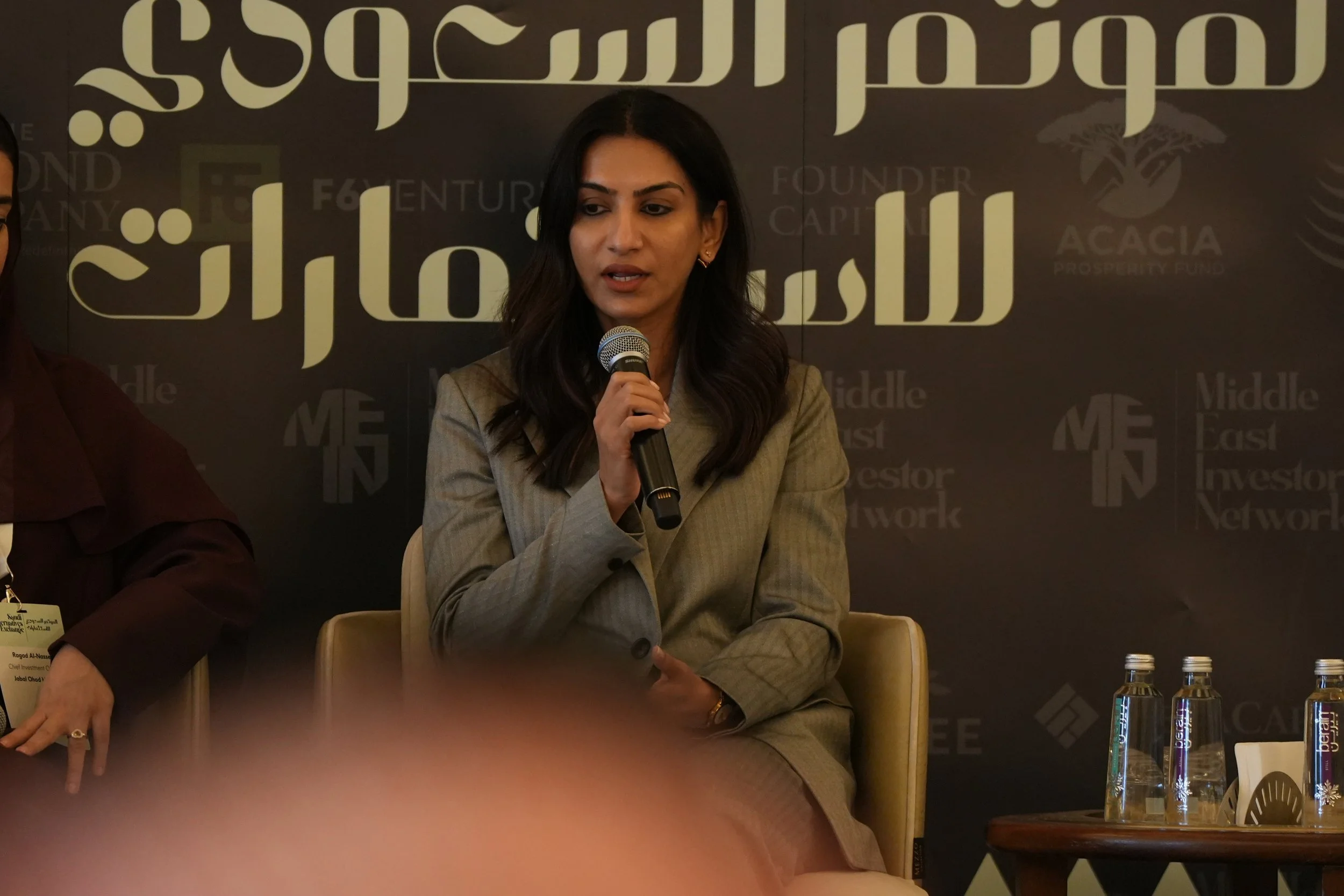 A woman holding a microphone and speaking at a panel discussion. She is dressed in a beige blazer and seated at a table with water bottles. The background features a dark wall with large Arabic-style script and various words like "founder," "capital,