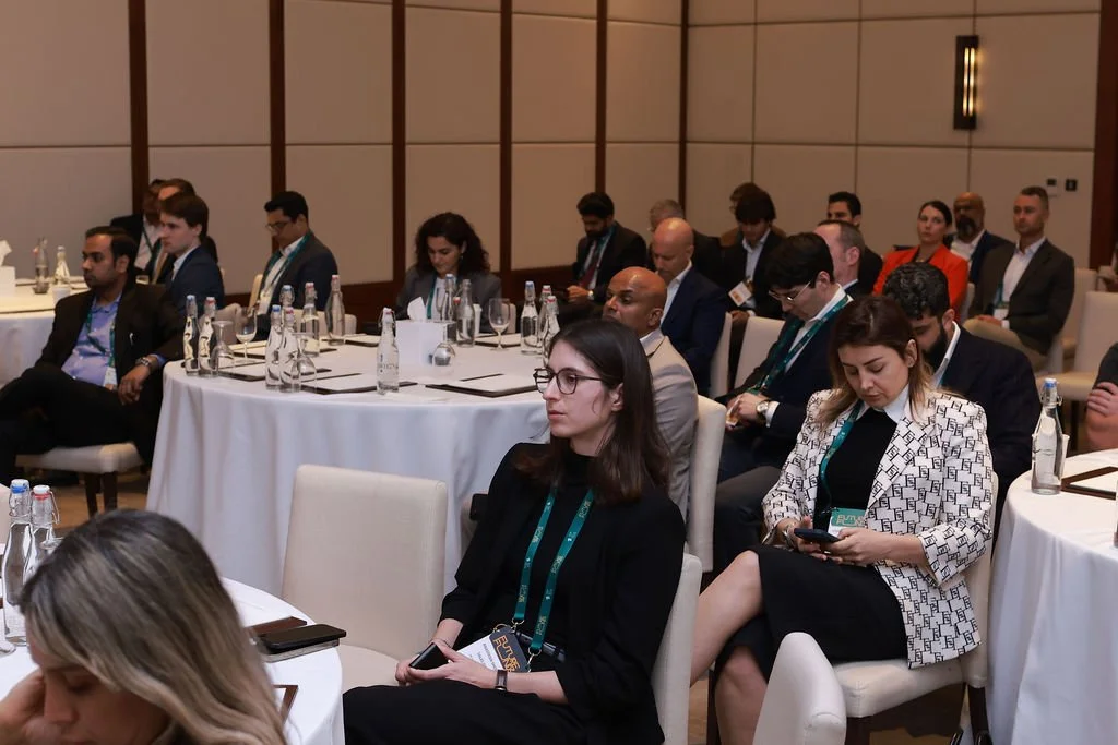Attendees seated at round tables in a conference room, some looking at devices, listening, or taking notes.