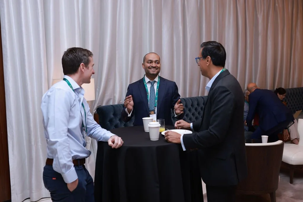 Three men in business attire are standing around a tall, round table, engaged in conversation and smiling. The setting appears to be a professional conference or networking event, with other attendees in the background.