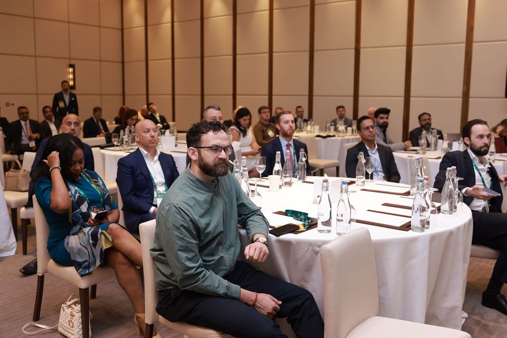 Business professionals attending a conference or seminar in a banquet hall, seated at round tables with water bottles, notepads, and pens.