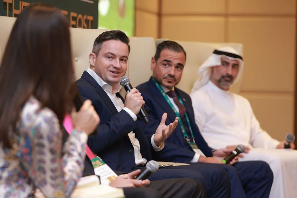 Four people sitting on a panel, three men and one woman. One man has a microphone and appears to be speaking, the others are holding microphones or listening. The setting is a conference or seminar.