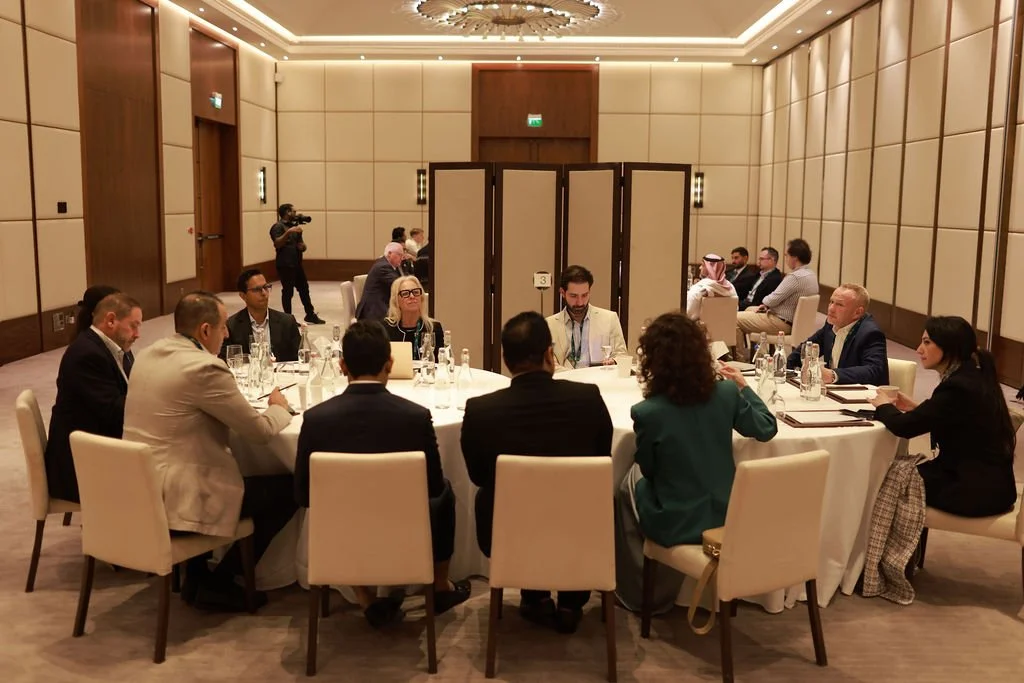 Business meeting with around 14 people seated around a round table in a luxury conference room, with three white display panels in the background and a camera operator filming the event.