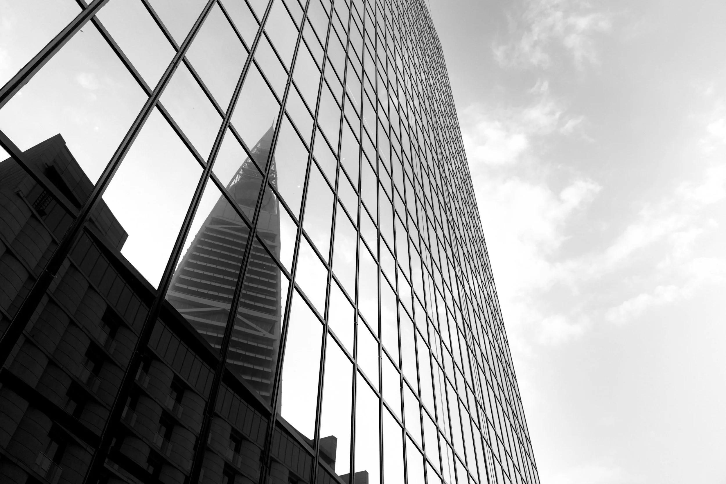 A low-angle view of a modern glass skyscraper reflecting the cloudy sky and nearby buildings.