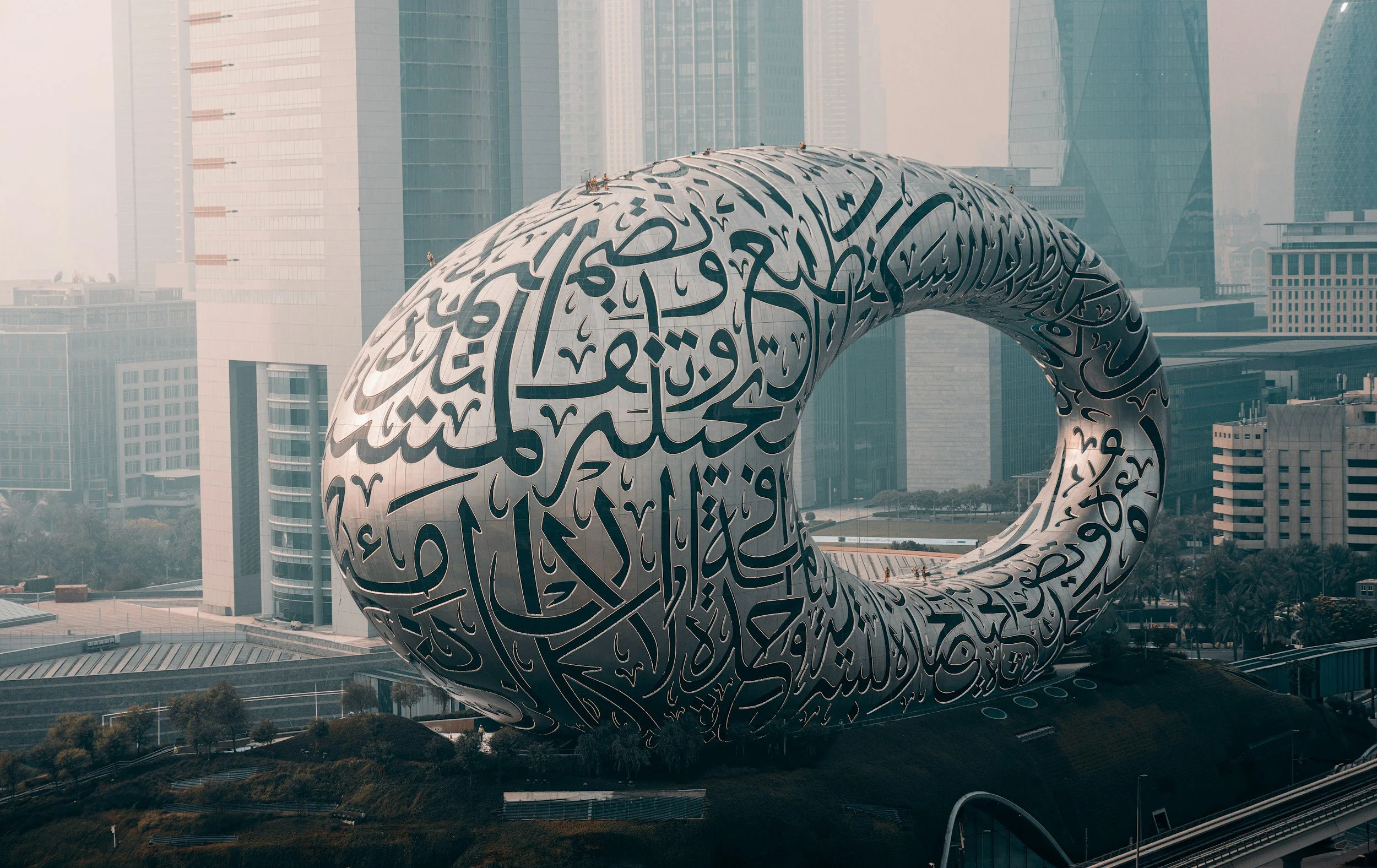 A large, modern sculpture with Arabic calligraphy, shaped like a circular loop on a cityscape background with tall glass buildings.