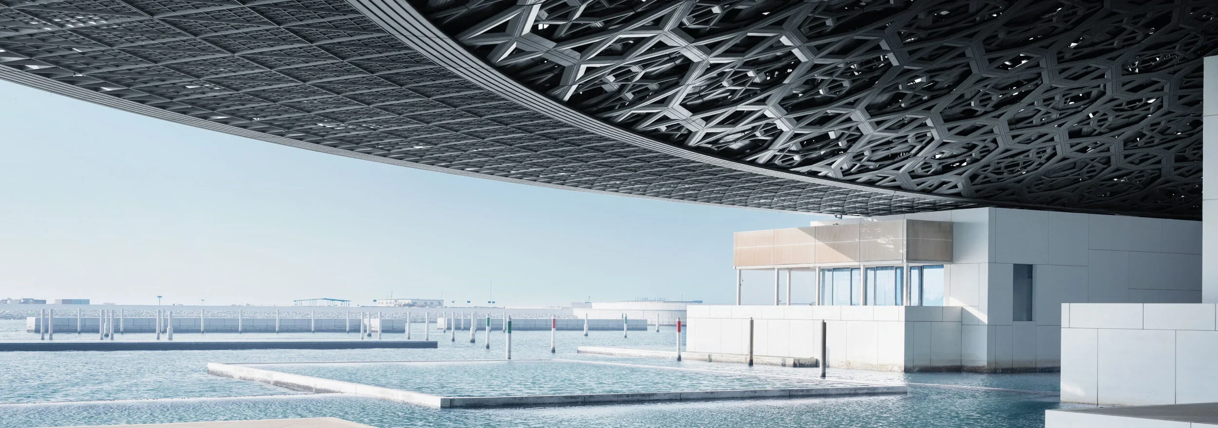 Modern architectural structure with a patterned metallic roof over a waterfront area. Clear sky and distant shoreline visible.