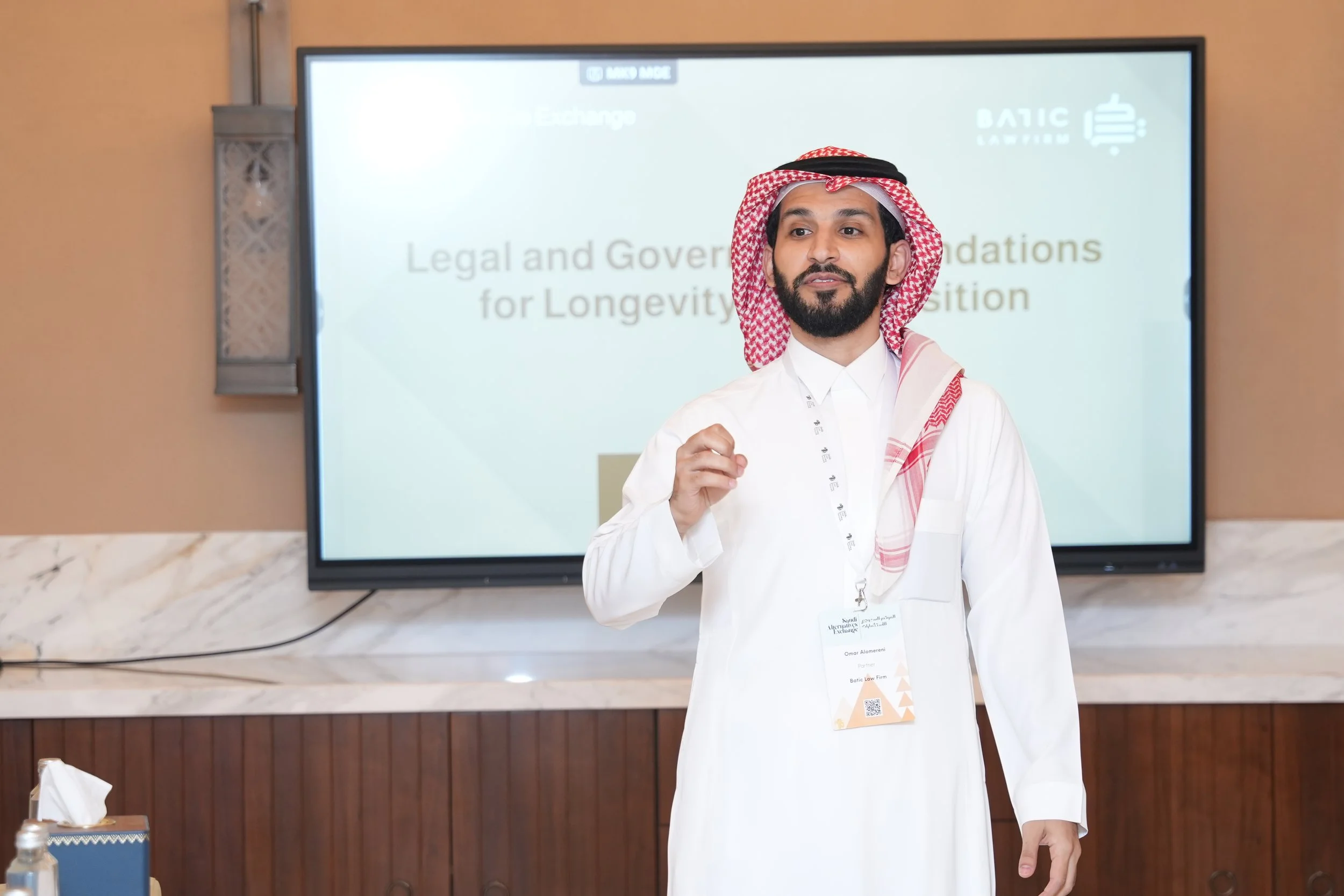 Man wearing traditional Middle Eastern attire, including a red and white checkered headscarf, standing in front of a presentation screen displaying a title about legal and governance foundations for longevity. The background includes a wall-mounted display, and there are items like hand sanitizer and tissues on a table in the foreground.