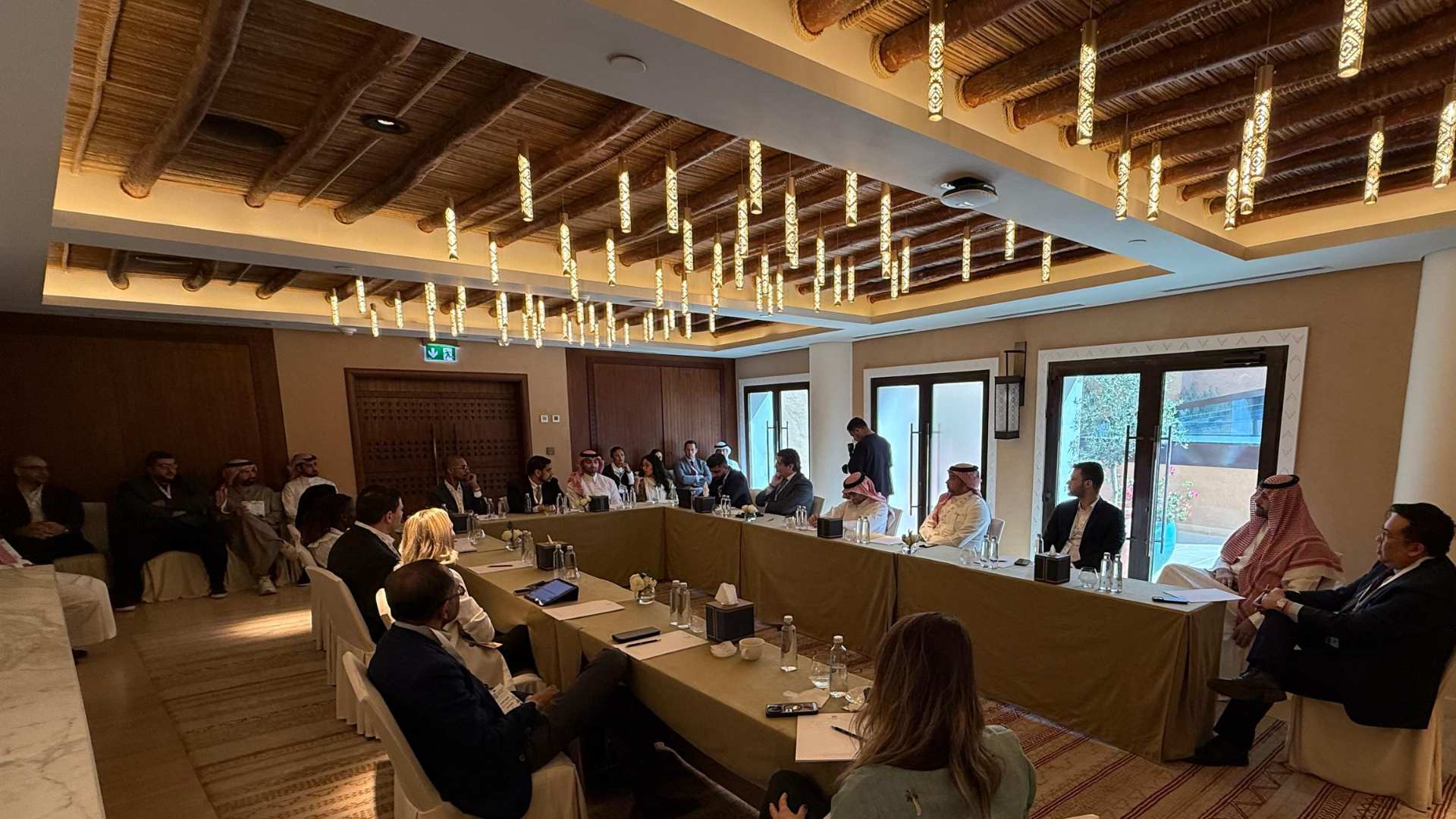 Business meeting or conference with diverse attendees seated around a large U-shaped table in a well-lit room with wooden accents and large windows.