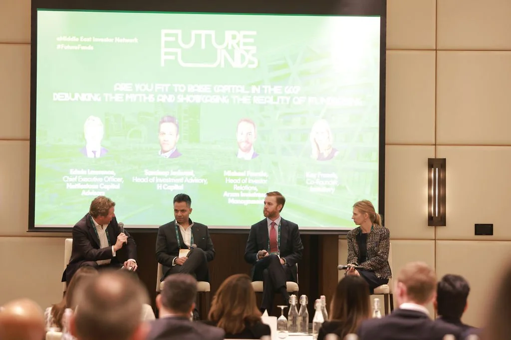 Panel discussion at the Middle East Investor Network conference with four speakers on stage, two women and two men, in front of a large green screen featuring the event logo and speaker profiles, with an audience in the foreground.